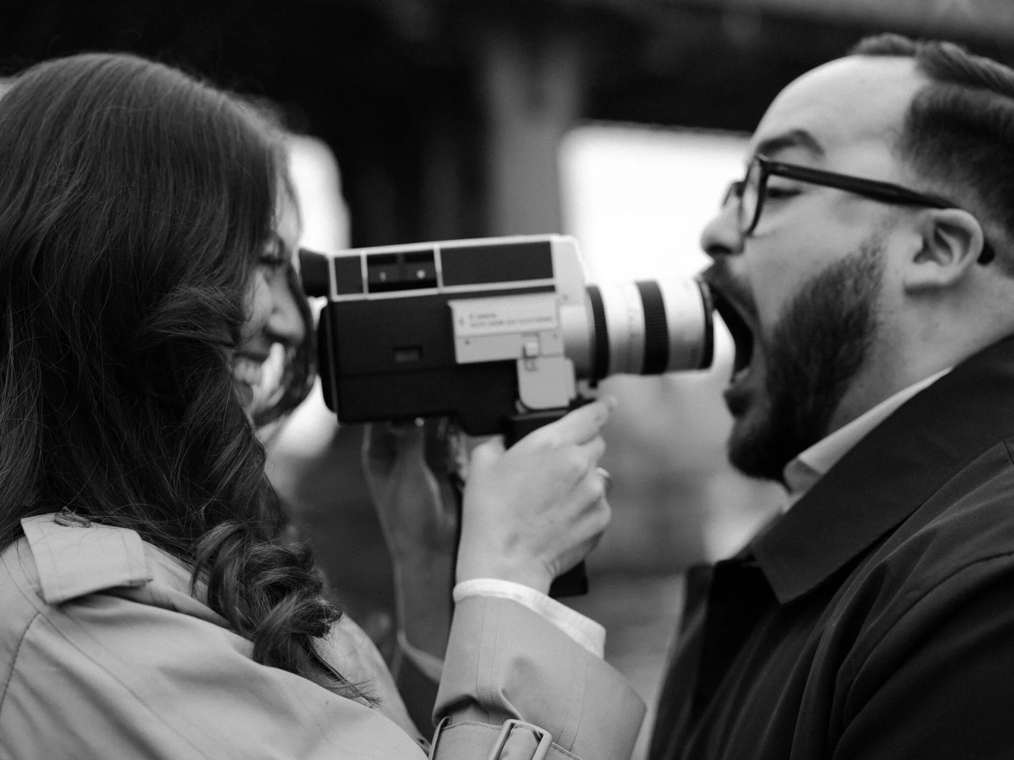 Playful black-and-white — Lexi points the Super-8 lens at Alfredo as if to feed him the camera; he opens his mouth wide in mock surprise.