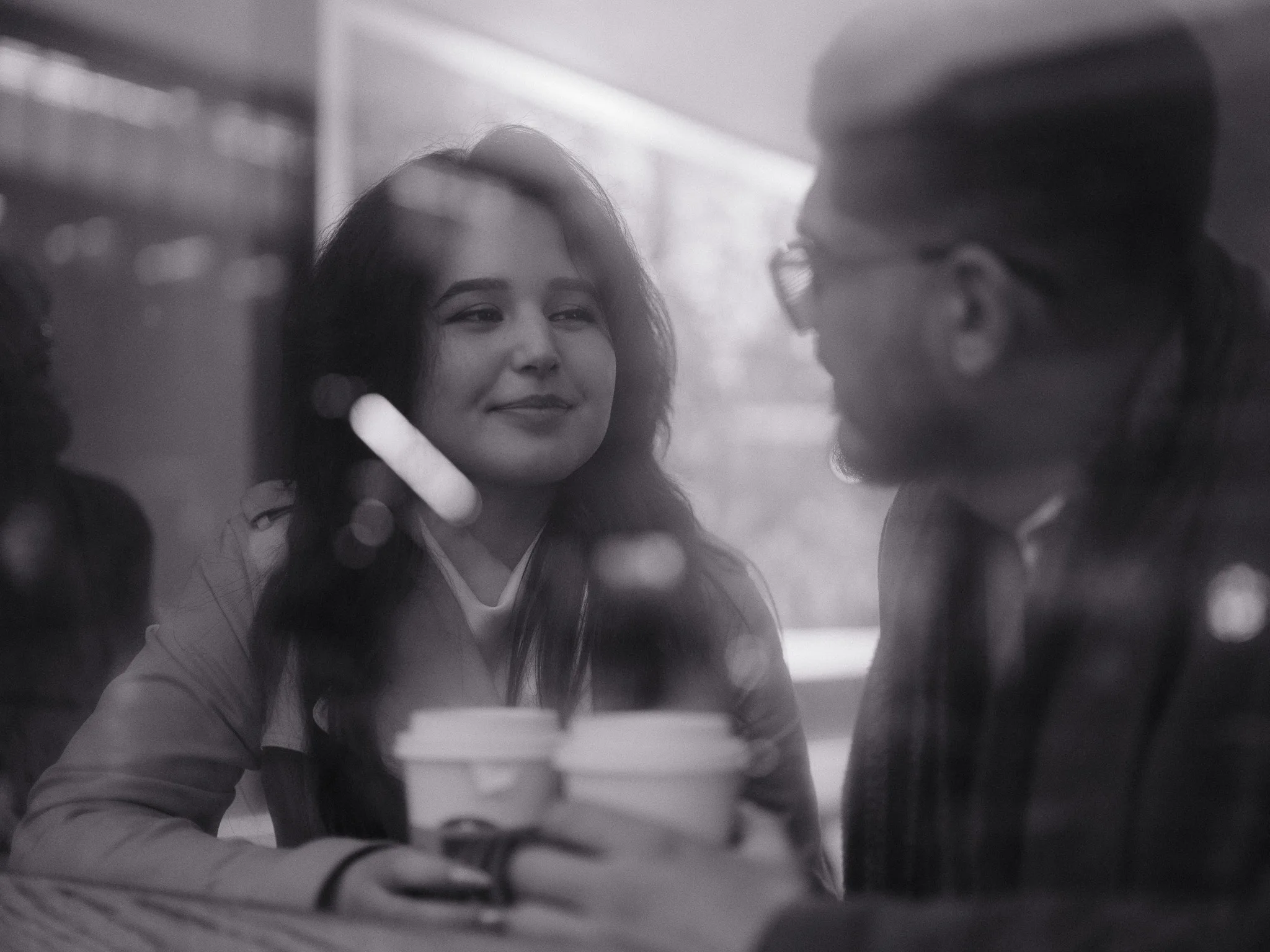 Through window glass at the diner — two coffees, Scarlett laughing at something Bayron said.