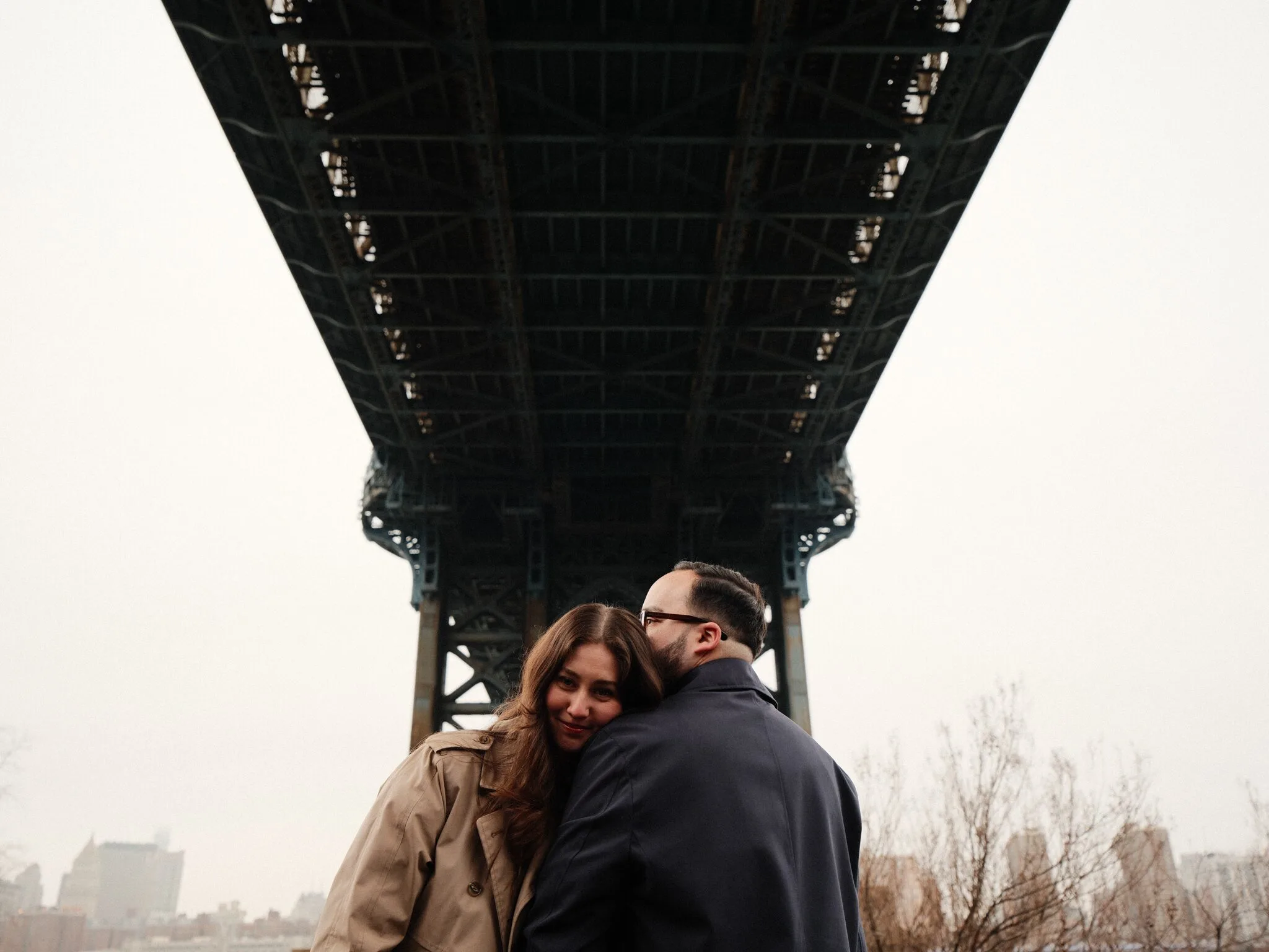 Black-and-white — a tight embrace under the bridge, Lexi's hair falling across Alfredo's shoulder, the chain-link fence blurred behind them.