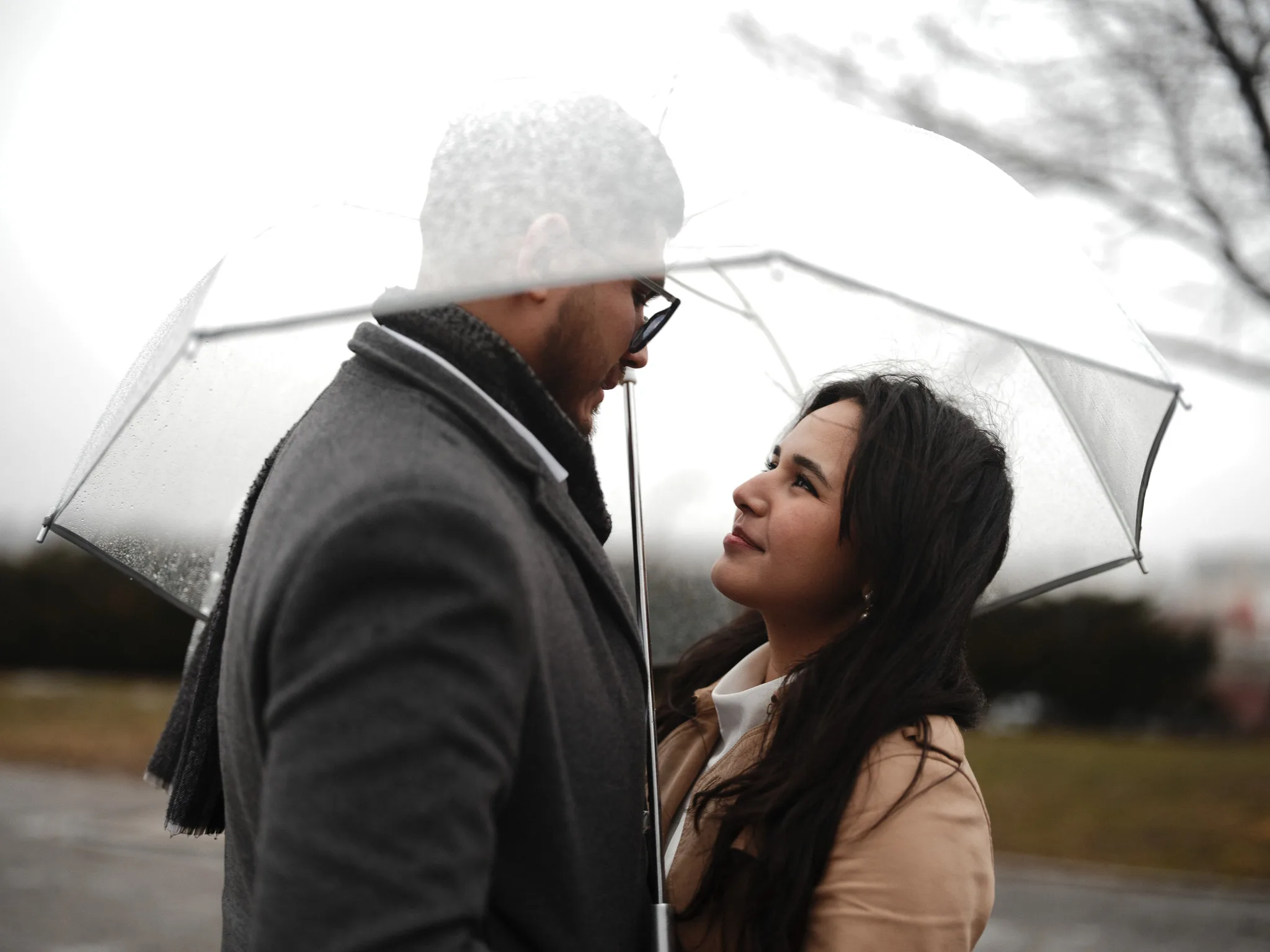 A couple under a clear umbrella on their wedding day — a wide exterior portrait in light rain.