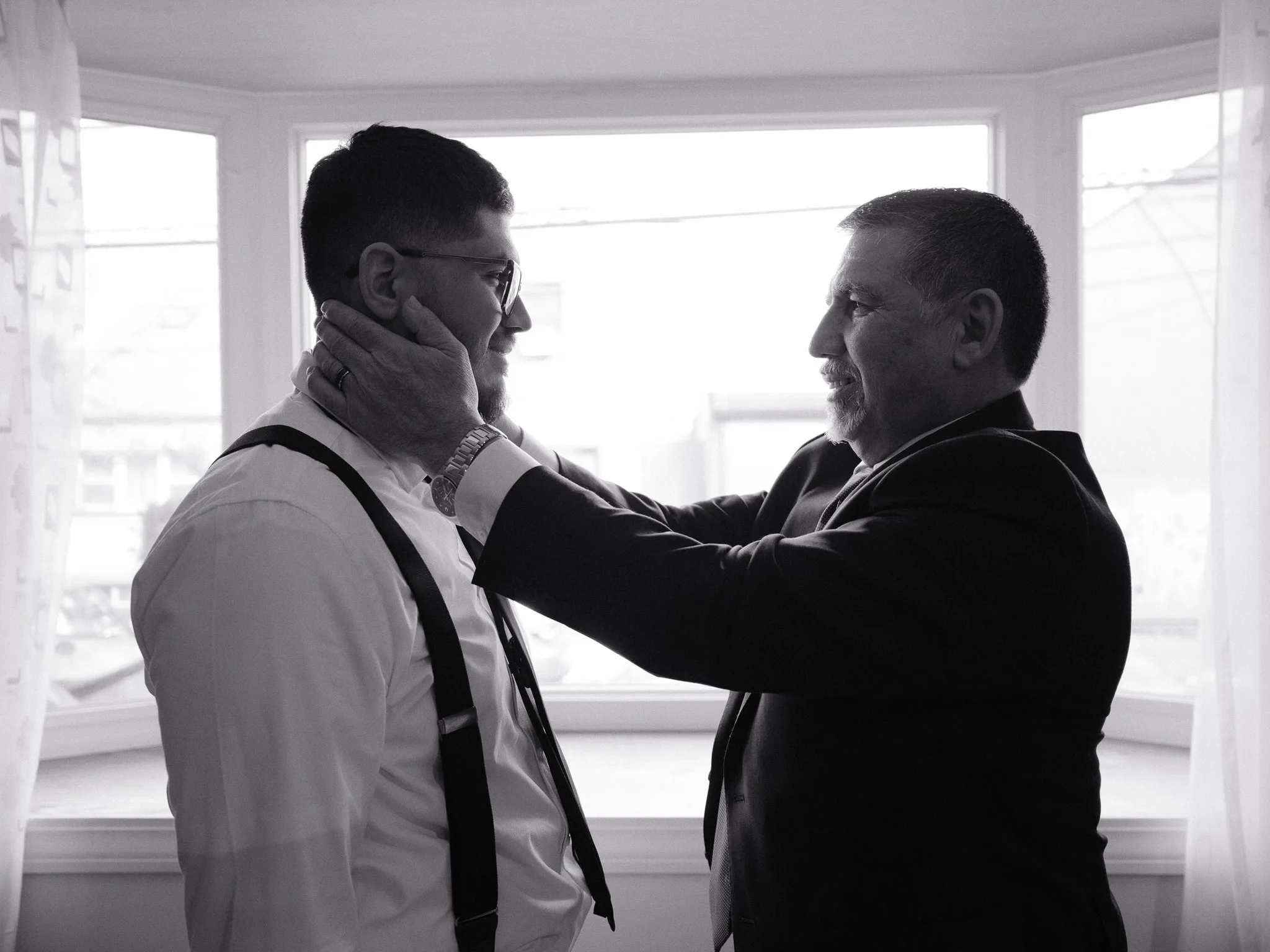 A father holds his son's face — Bayron in suspenders, white shirt, a window of light behind.