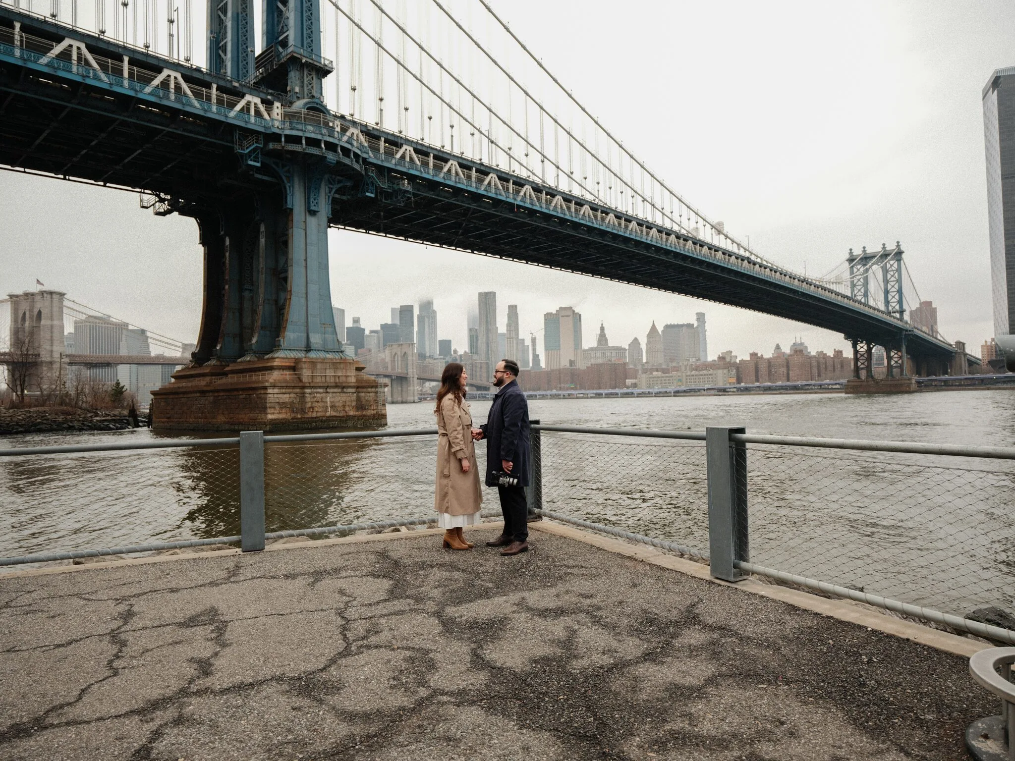 Couple standing together under the Manhattan Bridge with the New York City skyline in the background on an overcast day