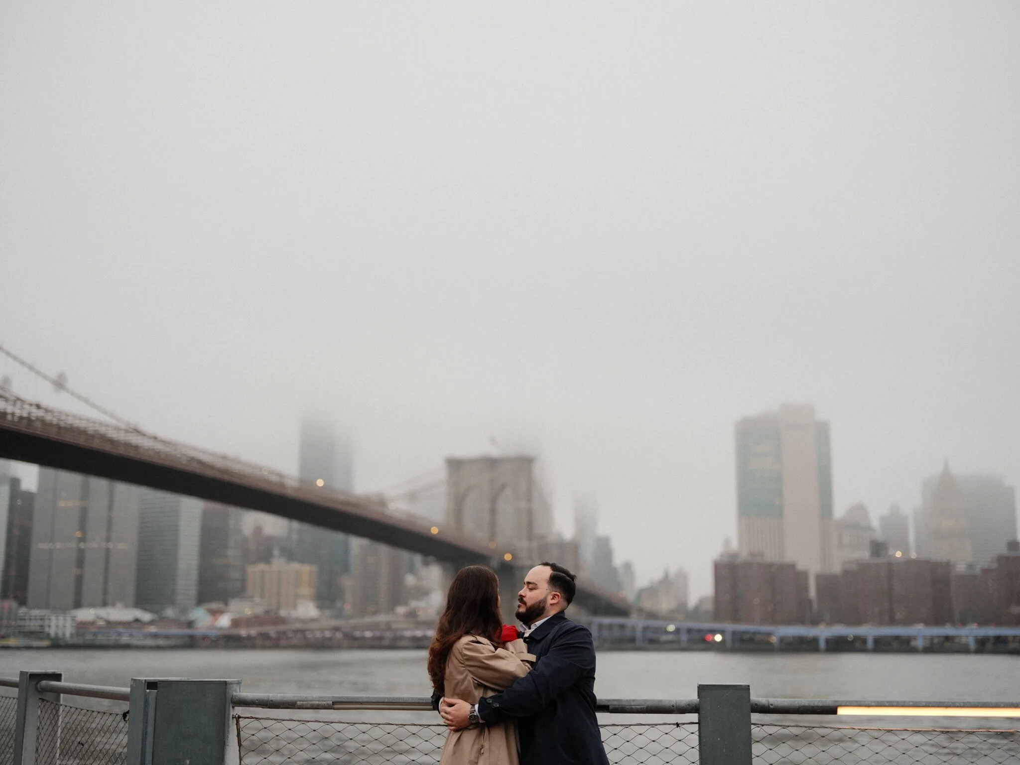 Alfredo and Lexi embrace on the Brooklyn waterfront; the Brooklyn Bridge and fog-shrouded Manhattan skyline rise behind them with a red rose between their coats.