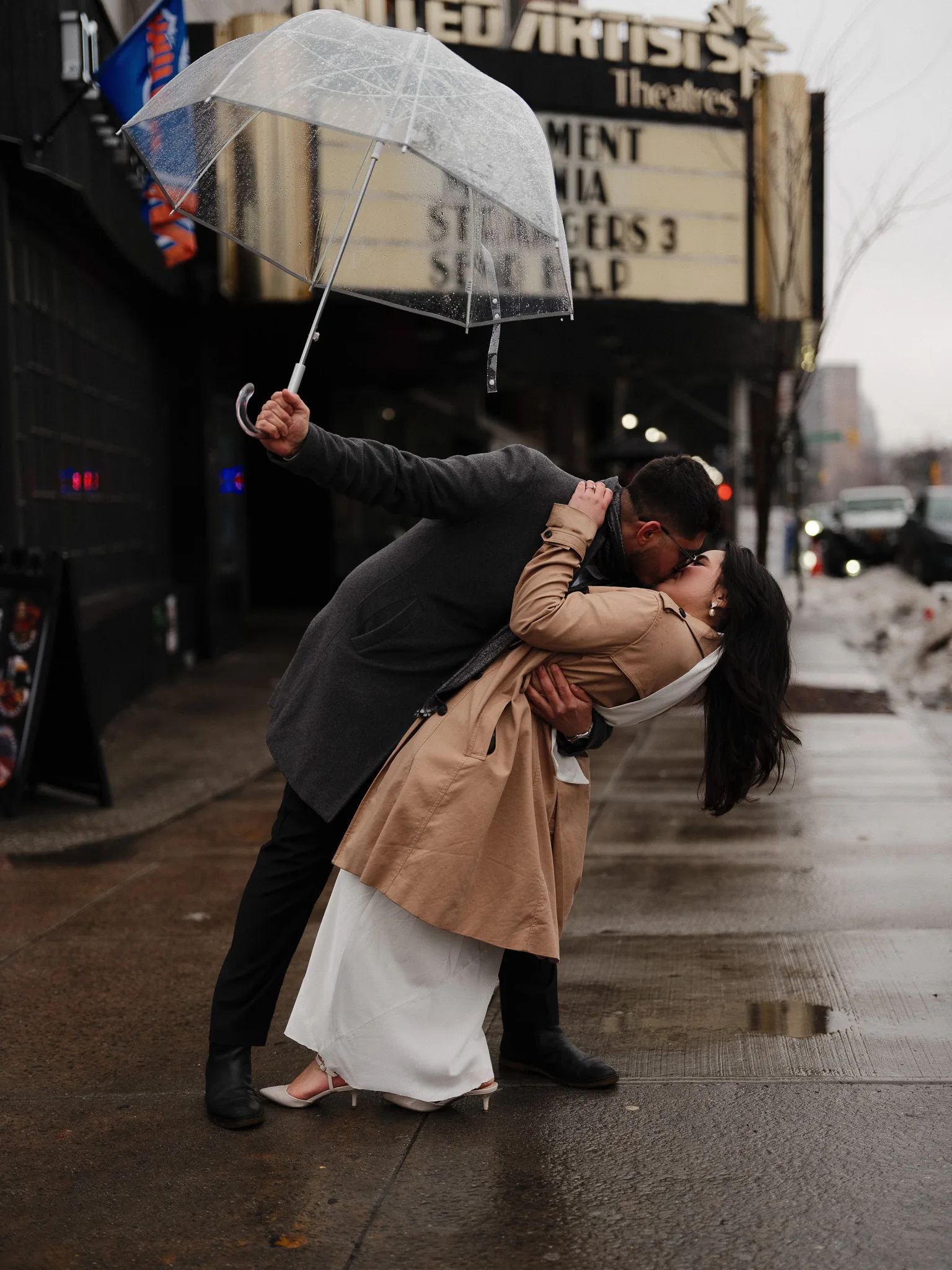 Couple sharing a dip kiss under a clear umbrella on a rainy city sidewalk outside a vintage theater marquee