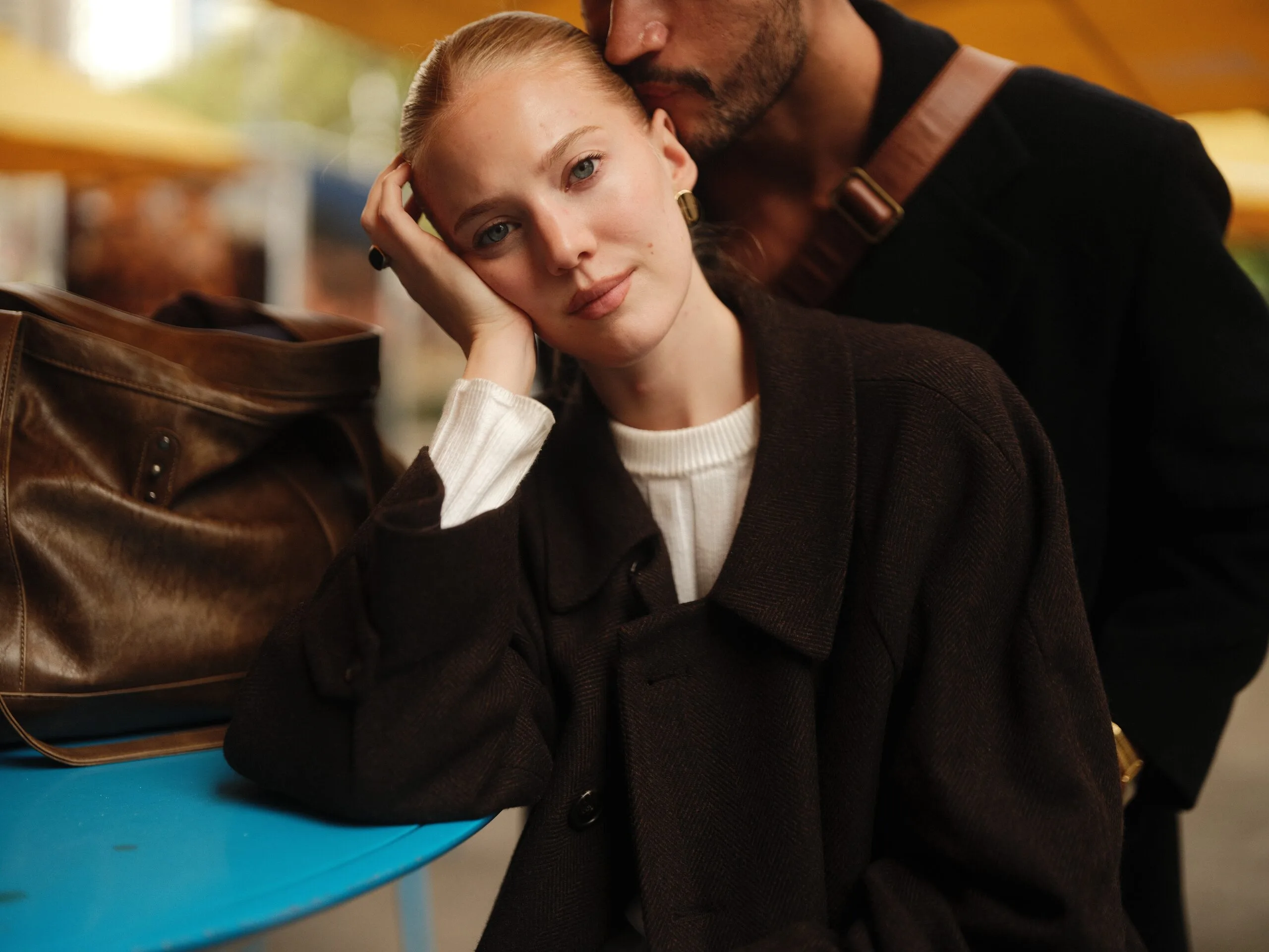 Rhylan leans into Juan at a café table, a brown leather bag beside them, his kiss at her temple.
