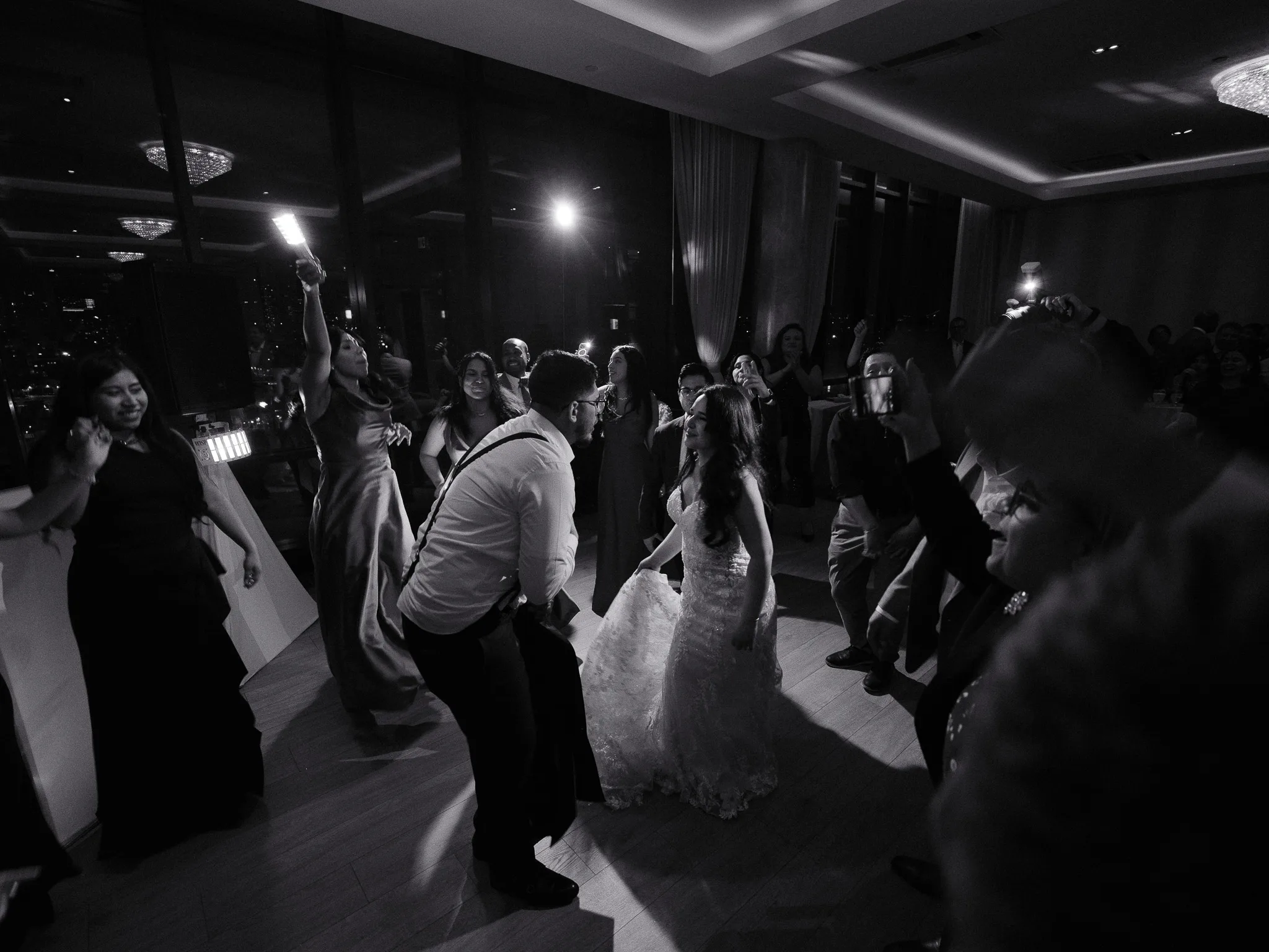 Bride and groom dancing together surrounded by guests on a lit wedding reception dance floor, black and white.