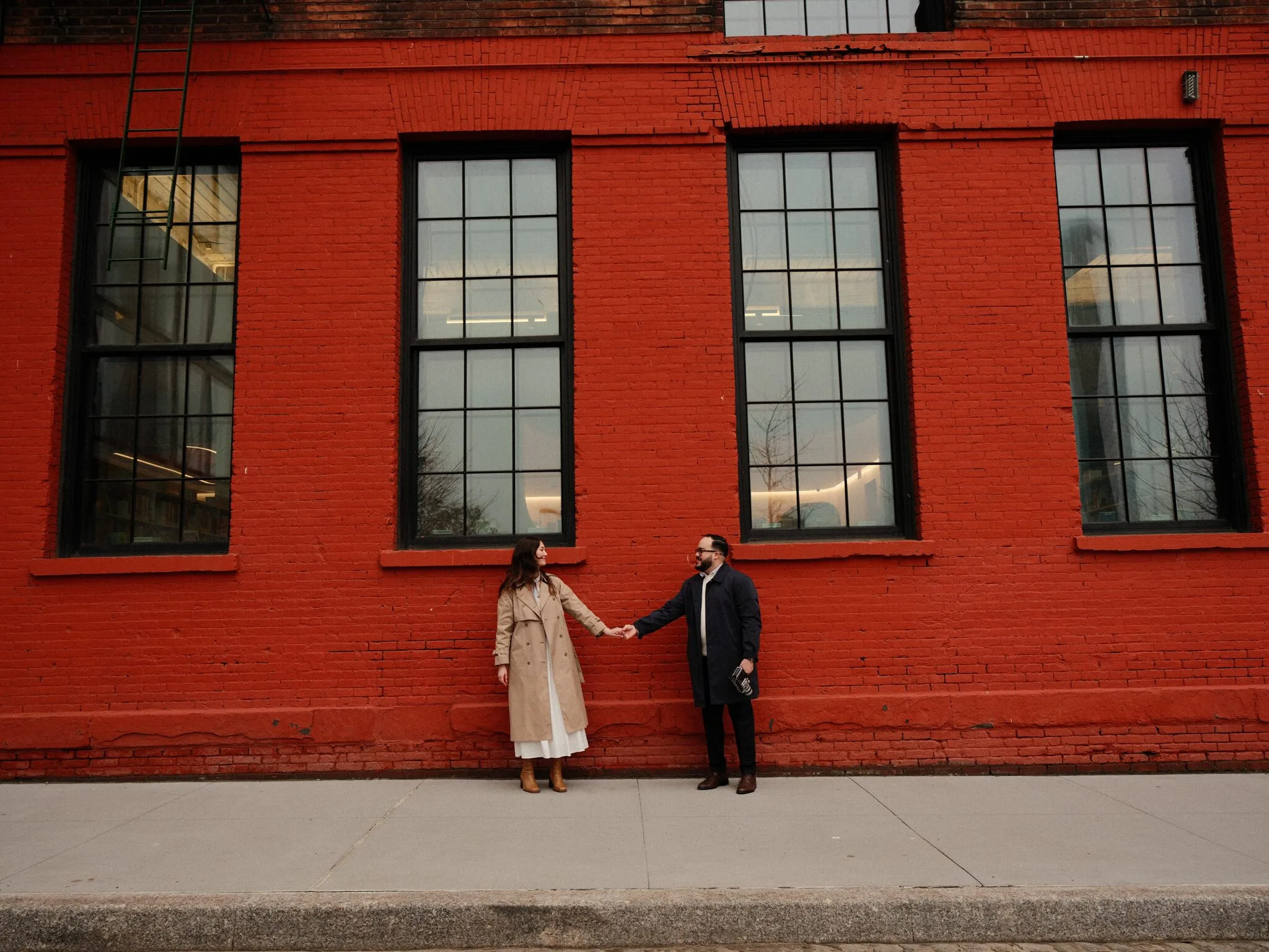 Alfredo and Lexi holding hands in front of a bold red-brick wall with three tall windows — a wide quiet composition, end of the day.