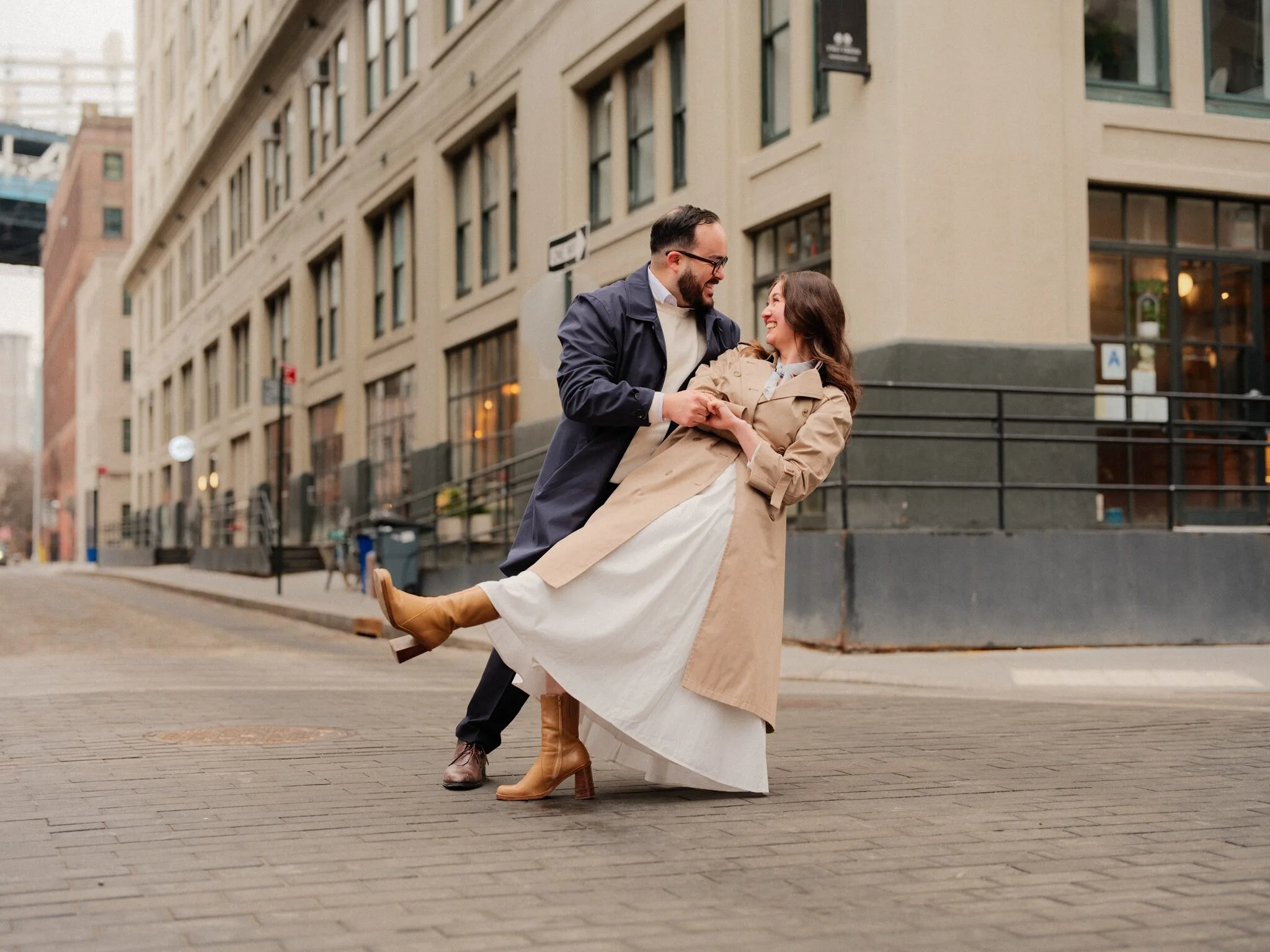 A dip on an empty cobblestone street — Alfredo holds Lexi mid-tip, her leg kicked out in a tan boot, both laughing; cream Dumbo buildings behind them.