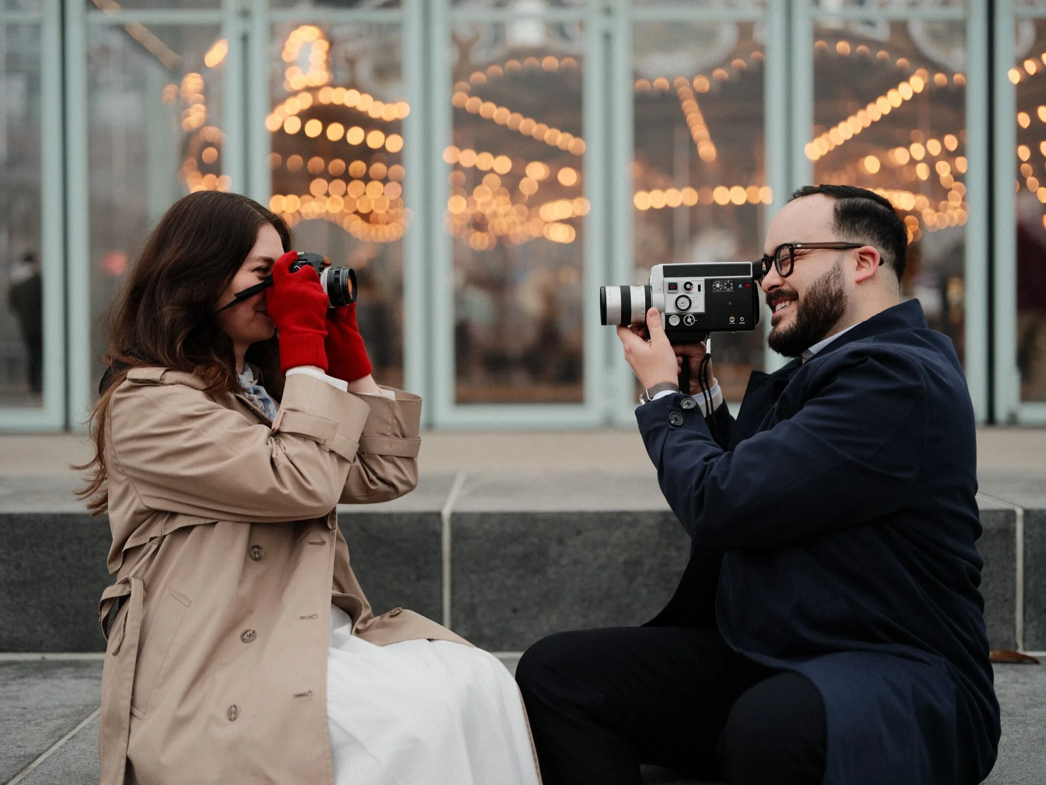 Lexi takes a photo with her small camera, red gloves bright against her beige trench coat; Alfredo lifts his Super-8 beside her, the carousel bulbs glowing behind them.