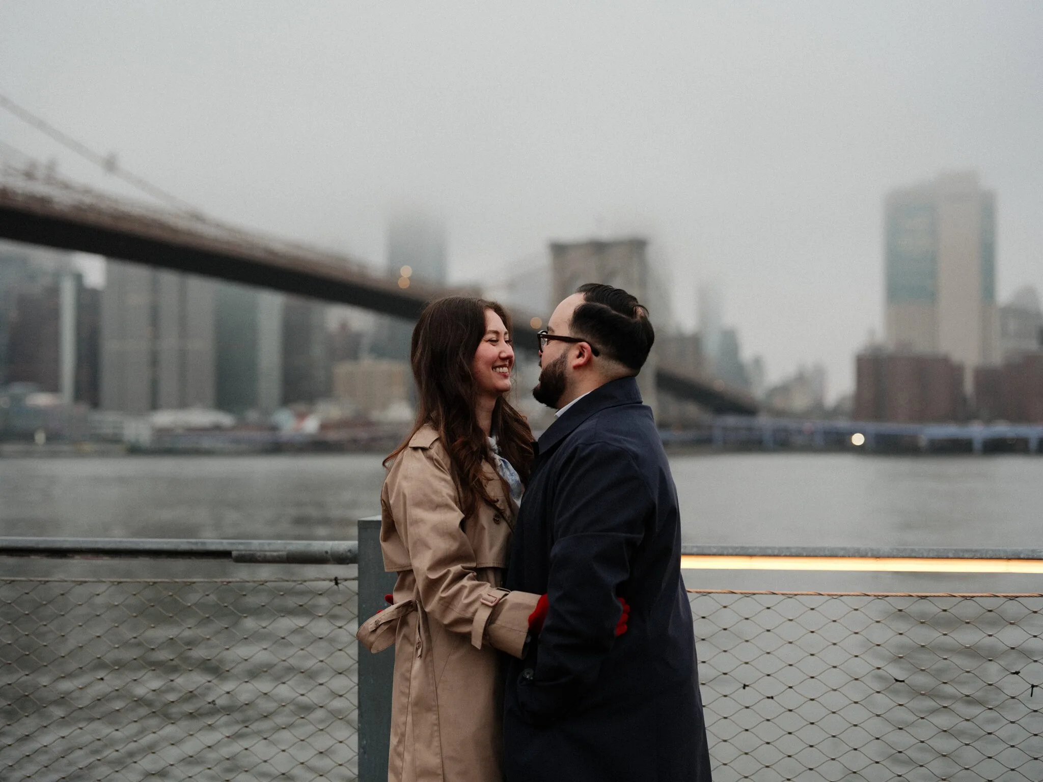 Alfredo and Lexi face each other at the waterfront rail, the Brooklyn Bridge curving into fog and the distant Manhattan skyline behind them.