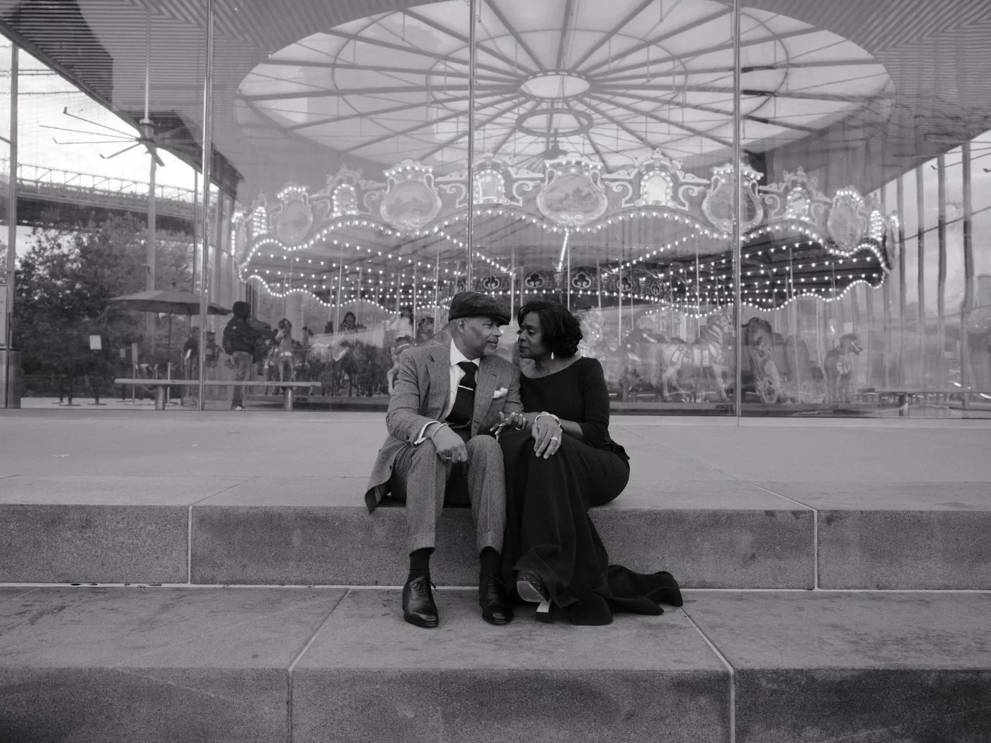 An elegant couple sitting close on a stone ledge in front of a carousel pavilion.