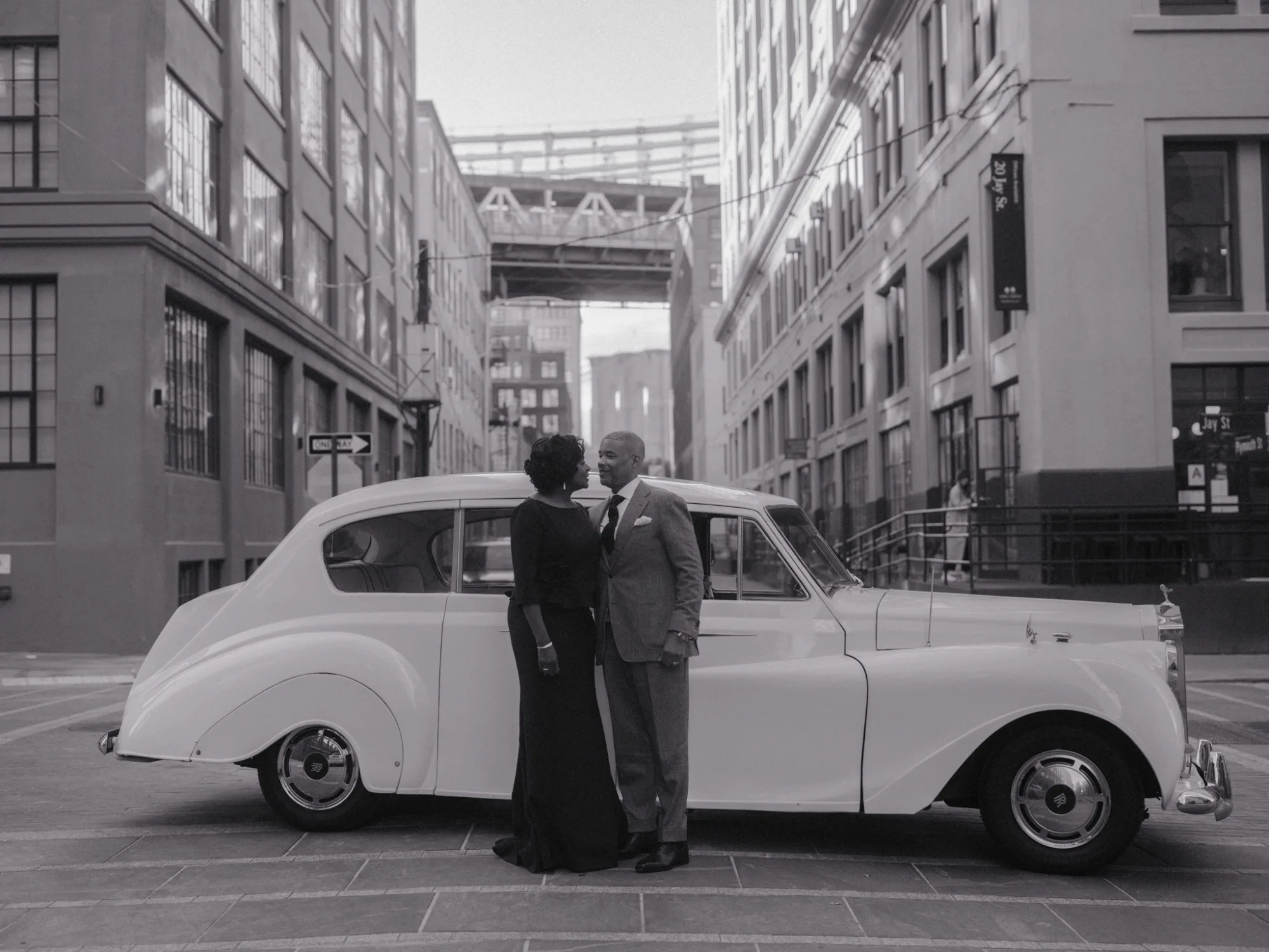 Marc and Olgeta walking hand in hand down a Dumbo street, gold afternoon light striping the brick behind them.