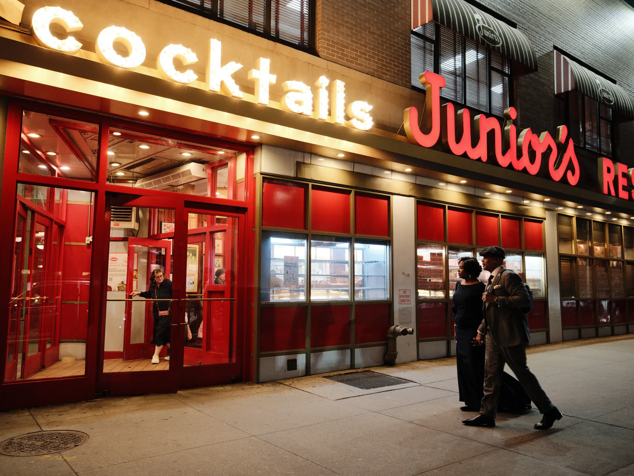 Marc and Olgeta arriving at Junior's diner on Flatbush Avenue at night, the iconic red sign and cocktails marquee glowing above them.