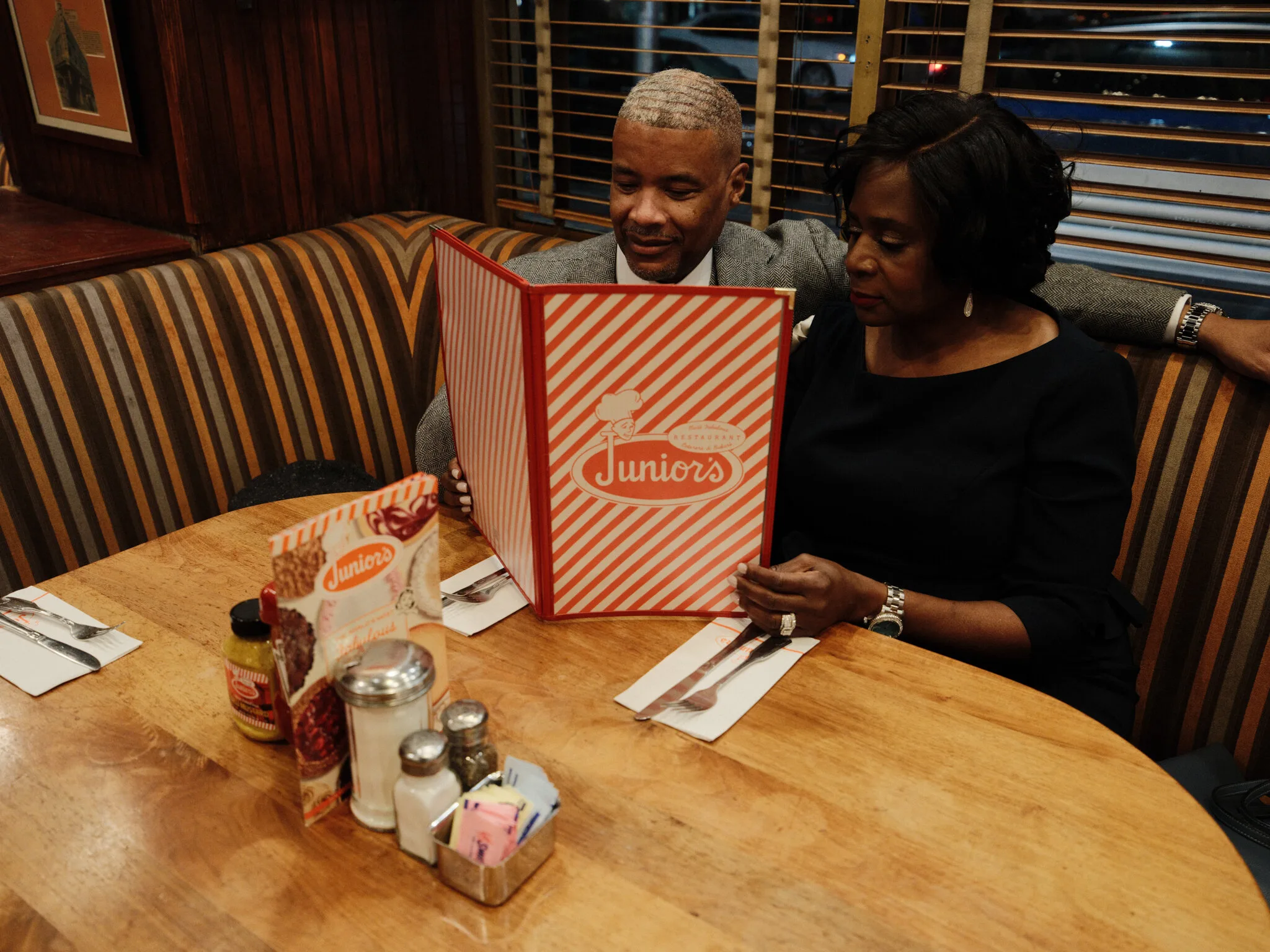 Marc and Olgeta seated together in a booth at Junior's, sharing a menu, warm diner light around them.