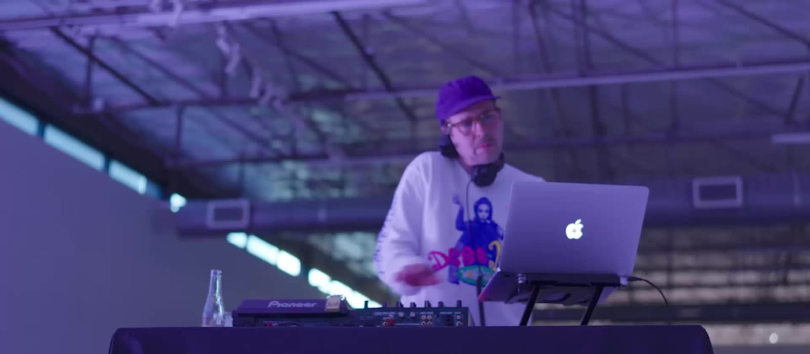A frame from a delivered reel: a DJ behind decks and a laptop under purple wash lighting in a large industrial event space
