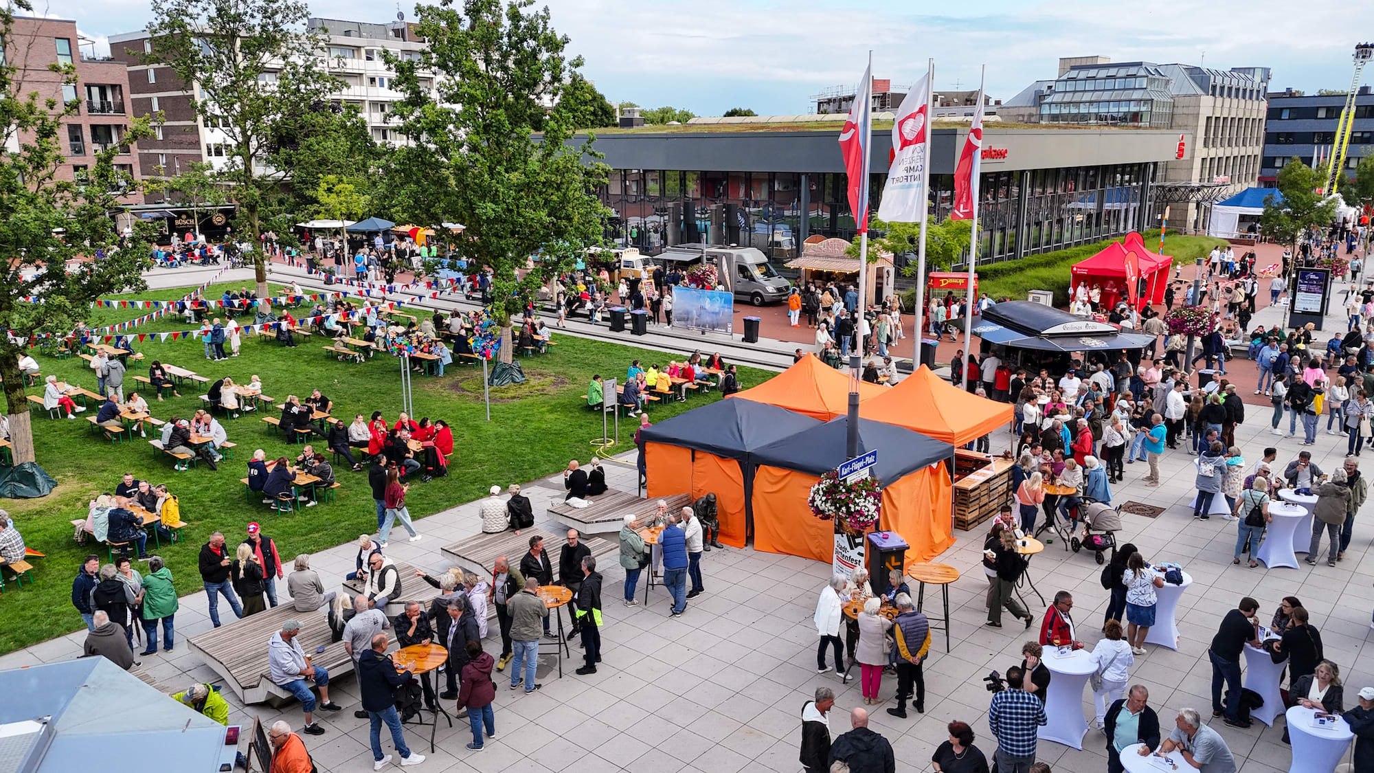 Luftaufnahme eines Stadtfestes auf dem Karl-Flügel-Platz in Kamp-Lintfort mit vielen Besuchern, bunten Pavillons, Bierbänken auf einer Grünfläche, Essensständen und dem Sparkassen-Gebäude im Hintergrund.