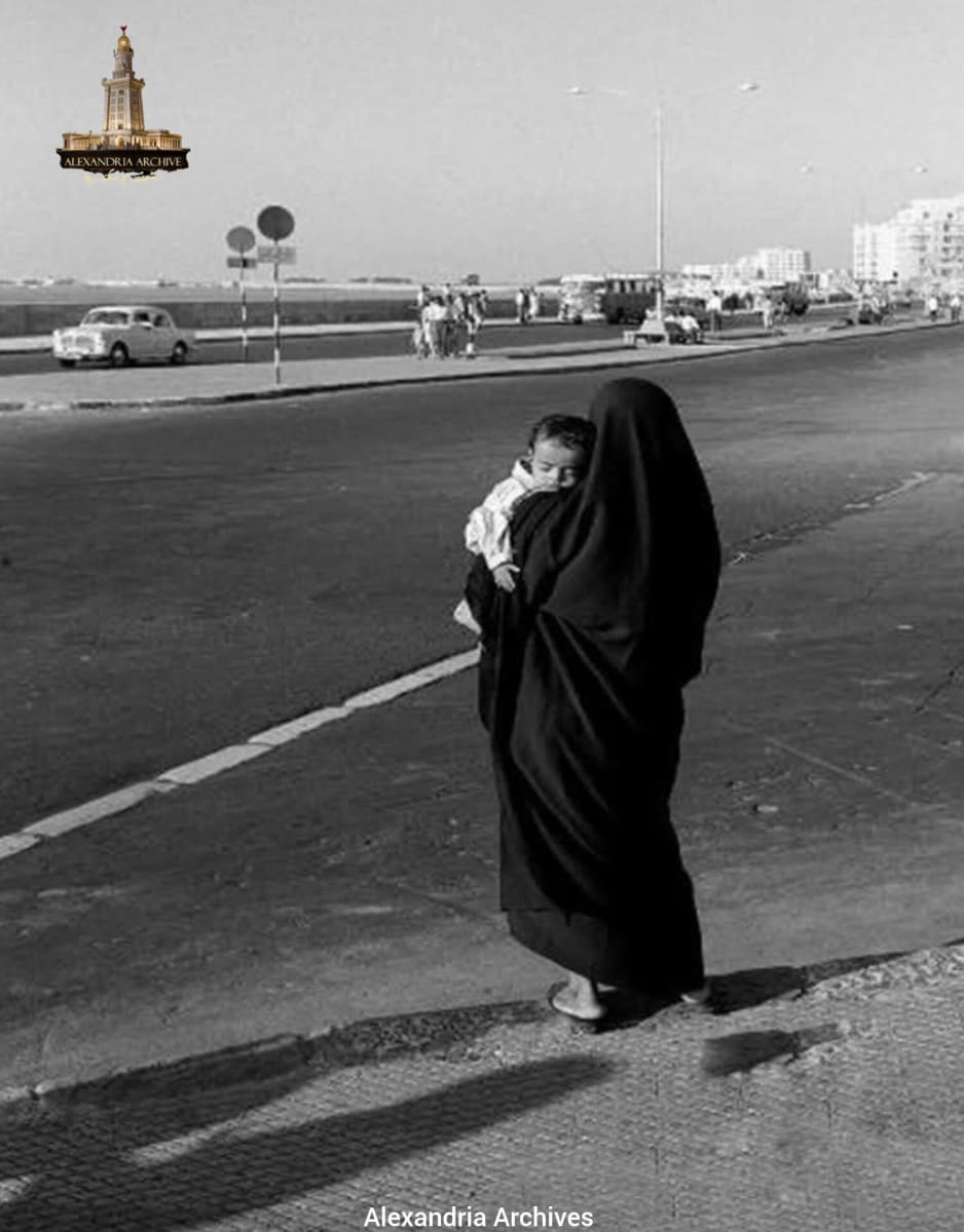 لقطة تاريخية توثق الهدوء على الكورنيش عام 1965تقربيا .  تظهر في الصورة سيدة اسكندرنية بالزي الشعبي التقليدي (الملاية اللف)  وفي الخلفية تظهر حركة النقل العام والسيارات الكلاسيكية التي ميزت شوارع عروس البحر المتوسط في منتصف الستينيات. 