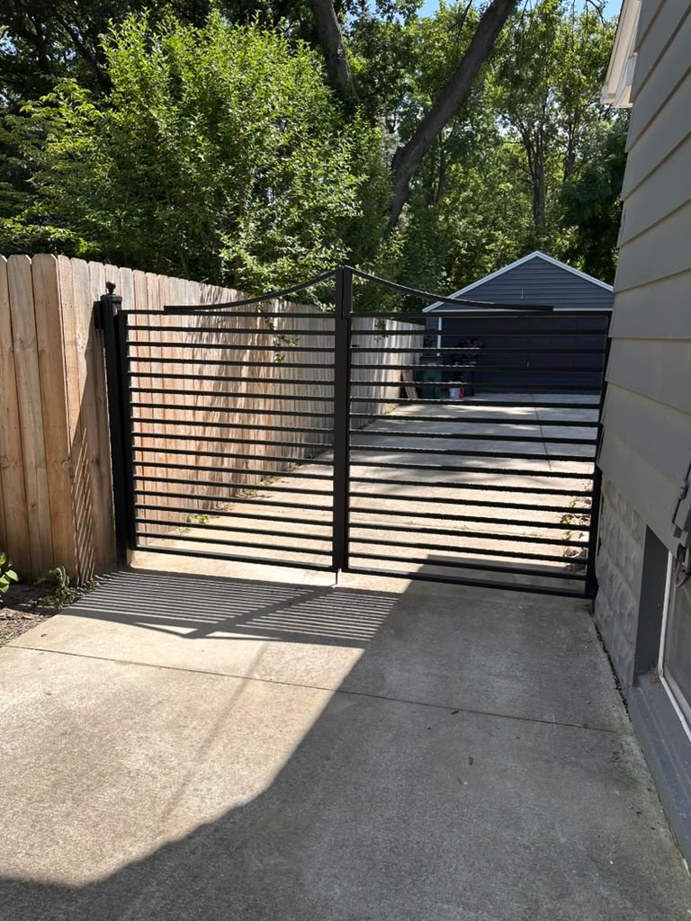 Custom black steel horizontal slat driveway gate installed between a wood fence and home in Warren, MI