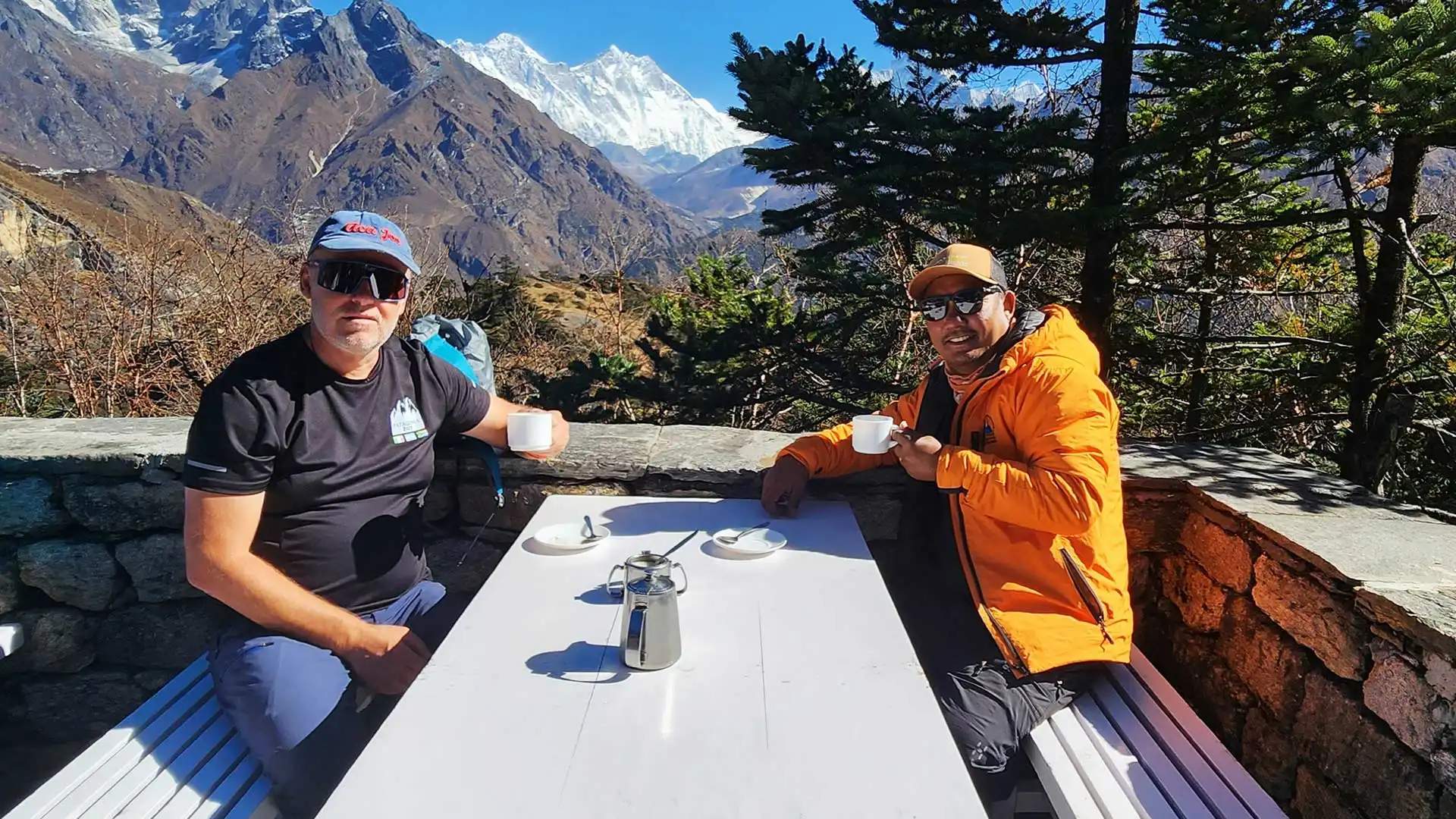 Two trekkers hold cups at an outdoor table with snowcapped mountains and pine trees behind.
