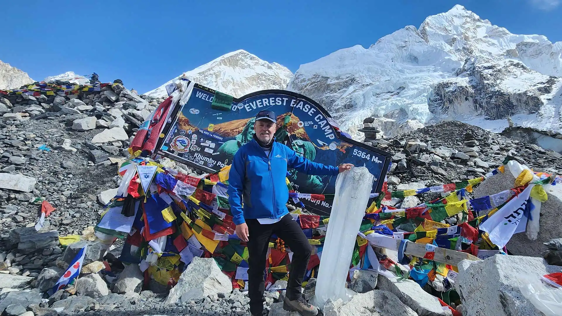 Trekker at Everest Base Camp beside colorful prayer flags, surrounded by rocky terrain and snowy peaks.