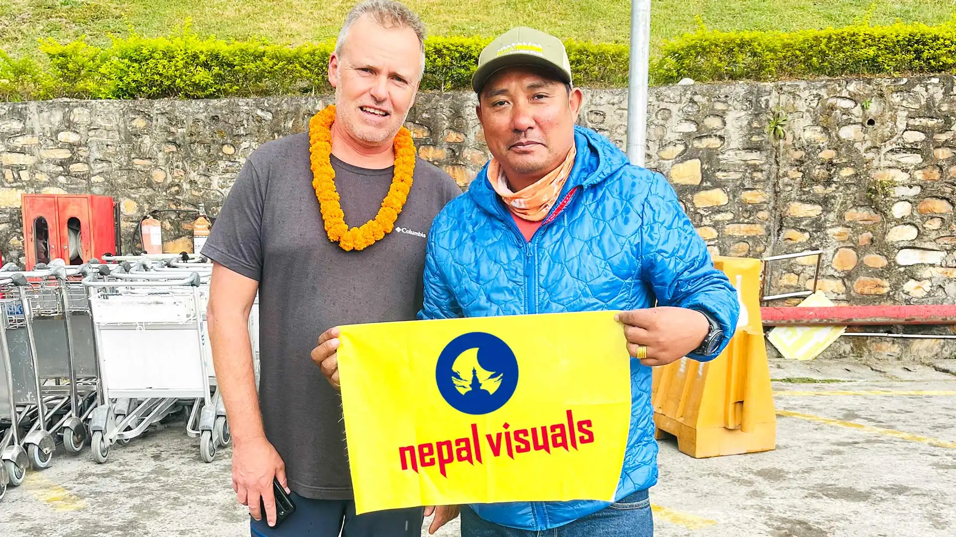 Two men holding a yellow Nepal Visuals banner, one wearing a marigold garland.