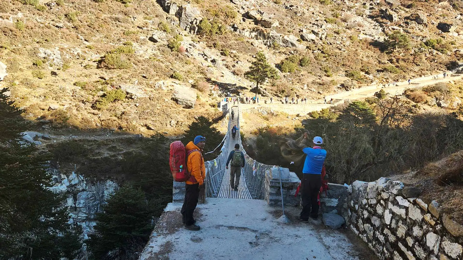 Trekkers crossing a narrow suspension bridge over a river in a rocky, forested valley.