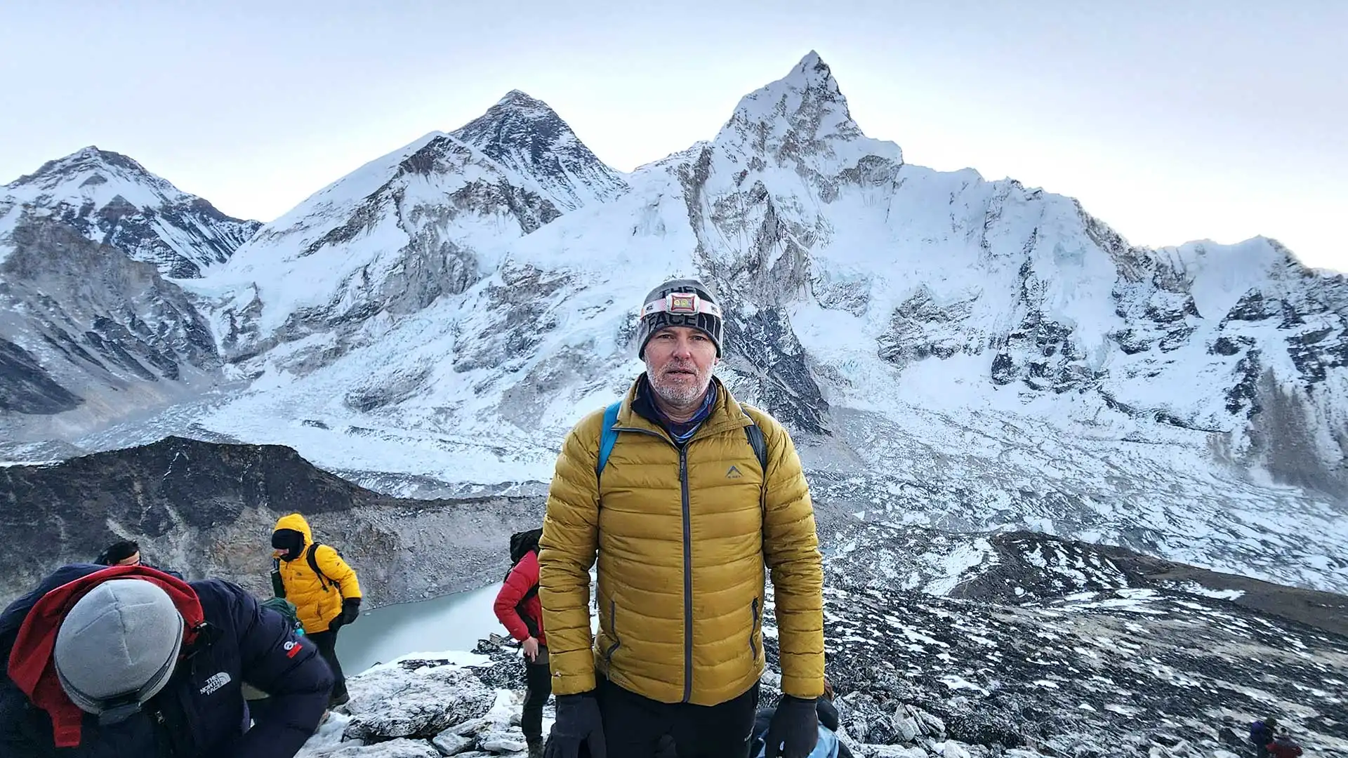 Trekker in a yellow jacket stands above a turquoise lake beneath snow-covered Himalayan peaks.
