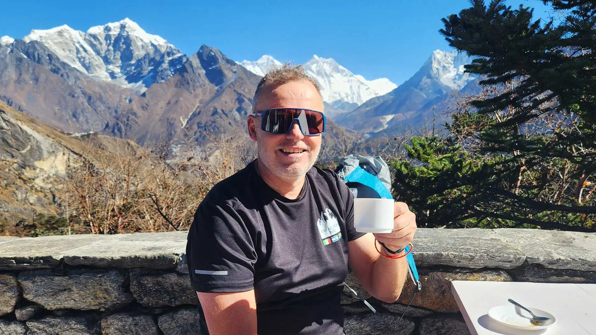Trekker wearing sunglasses holds a white mug with snowcapped mountains and pine trees behind him.