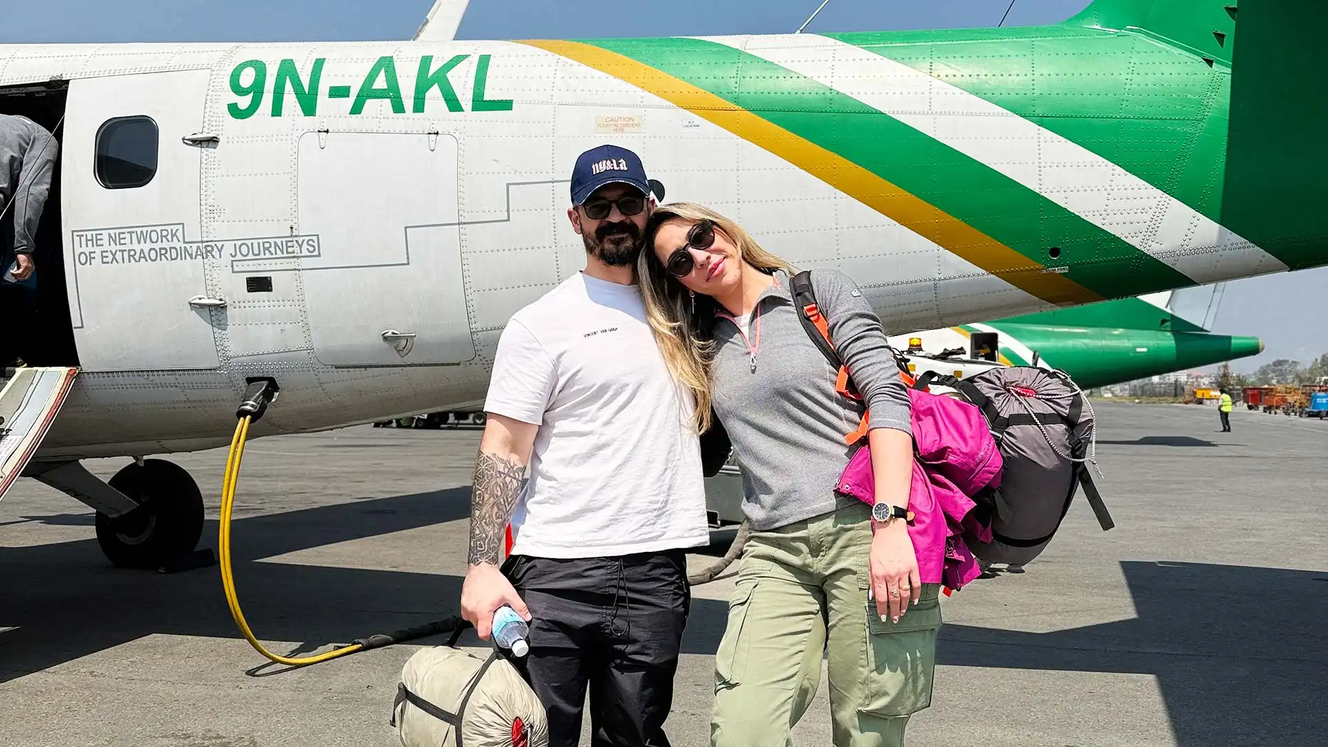 Two travelers with bags pose beside a small aircraft on an airport tarmac.
