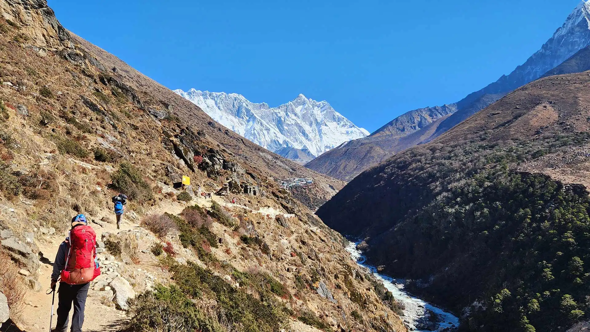 Trekkers follow a rocky mountainside trail above a river, with snow-covered peaks under clear skies.