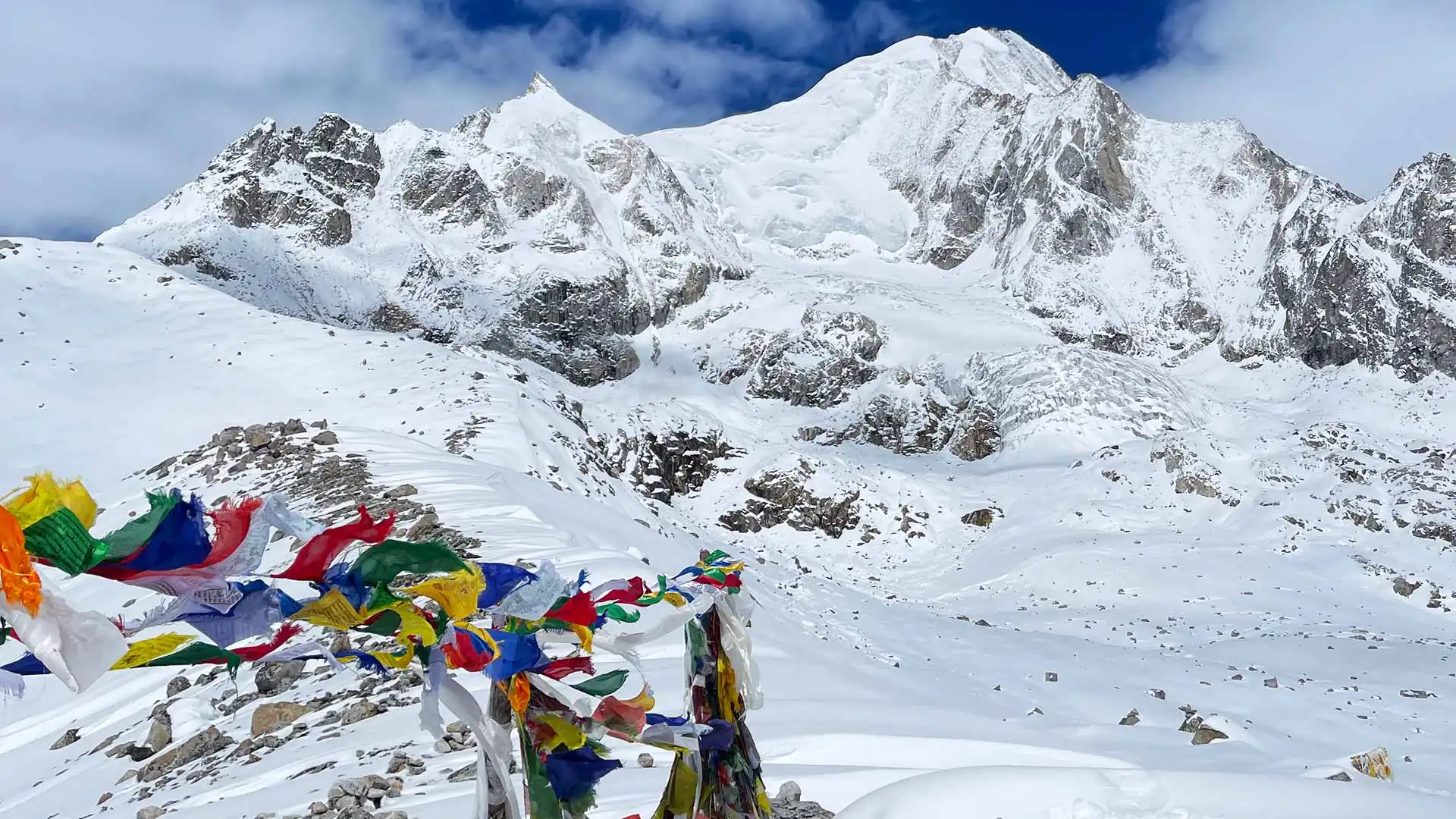 Colorful prayer flags flutter before snow-covered Himalayan peaks beneath a partly cloudy sky.