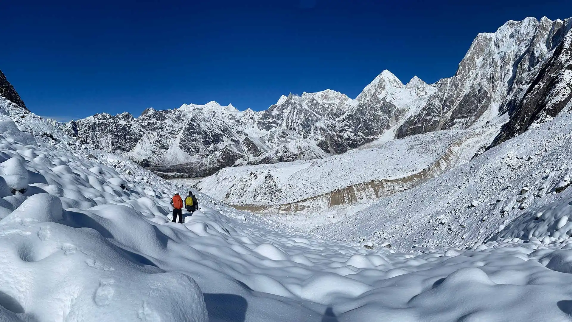 Two trekkers cross a snowy mountain valley beneath jagged peaks and clear blue skies.