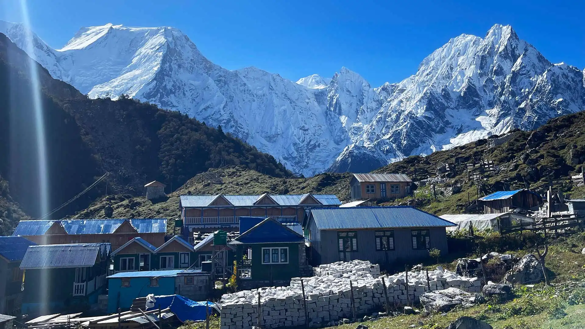 Stone houses with blue metal roofs sit beneath towering snow-covered mountains and clear skies.