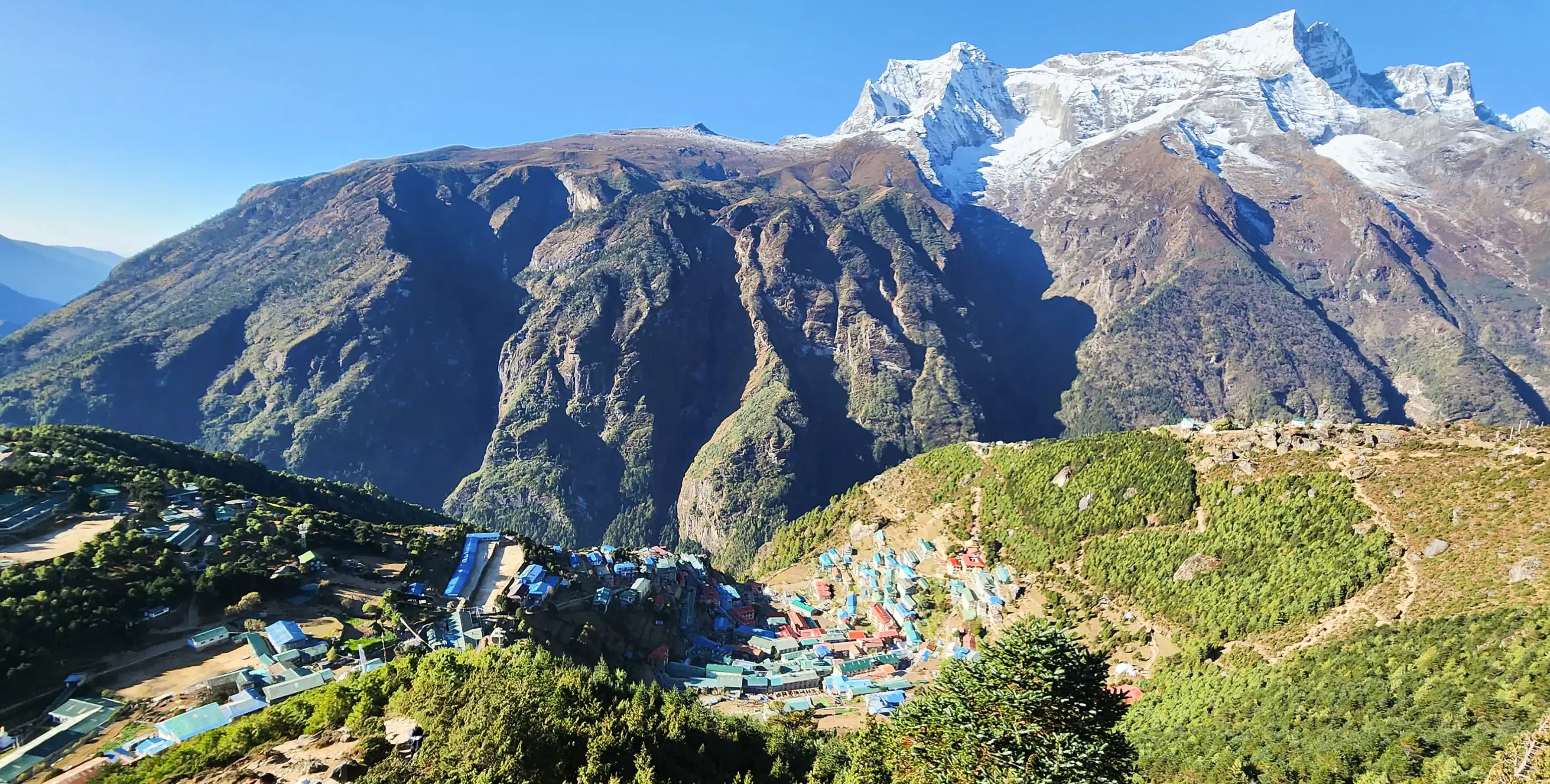 Colorful village buildings nestled among green hills beneath rugged, snow-capped mountains and clear blue skies.