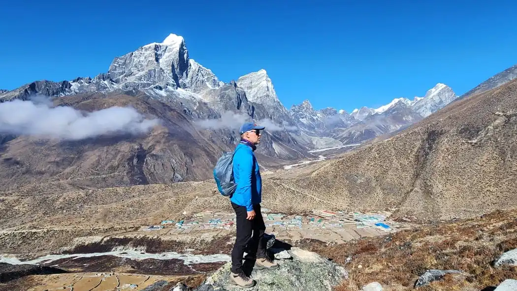 Trekker with backpack stands on a rocky overlook above a village beneath snow-dusted Himalayan peaks.