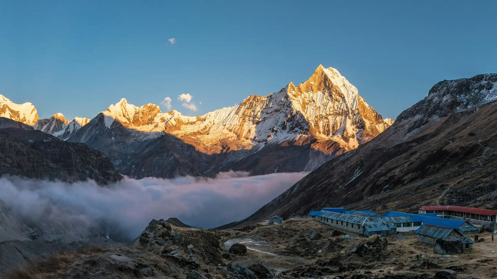 Golden sunlight illuminates snowy peaks above a cloud-filled valley and blue-roofed trekking lodges.