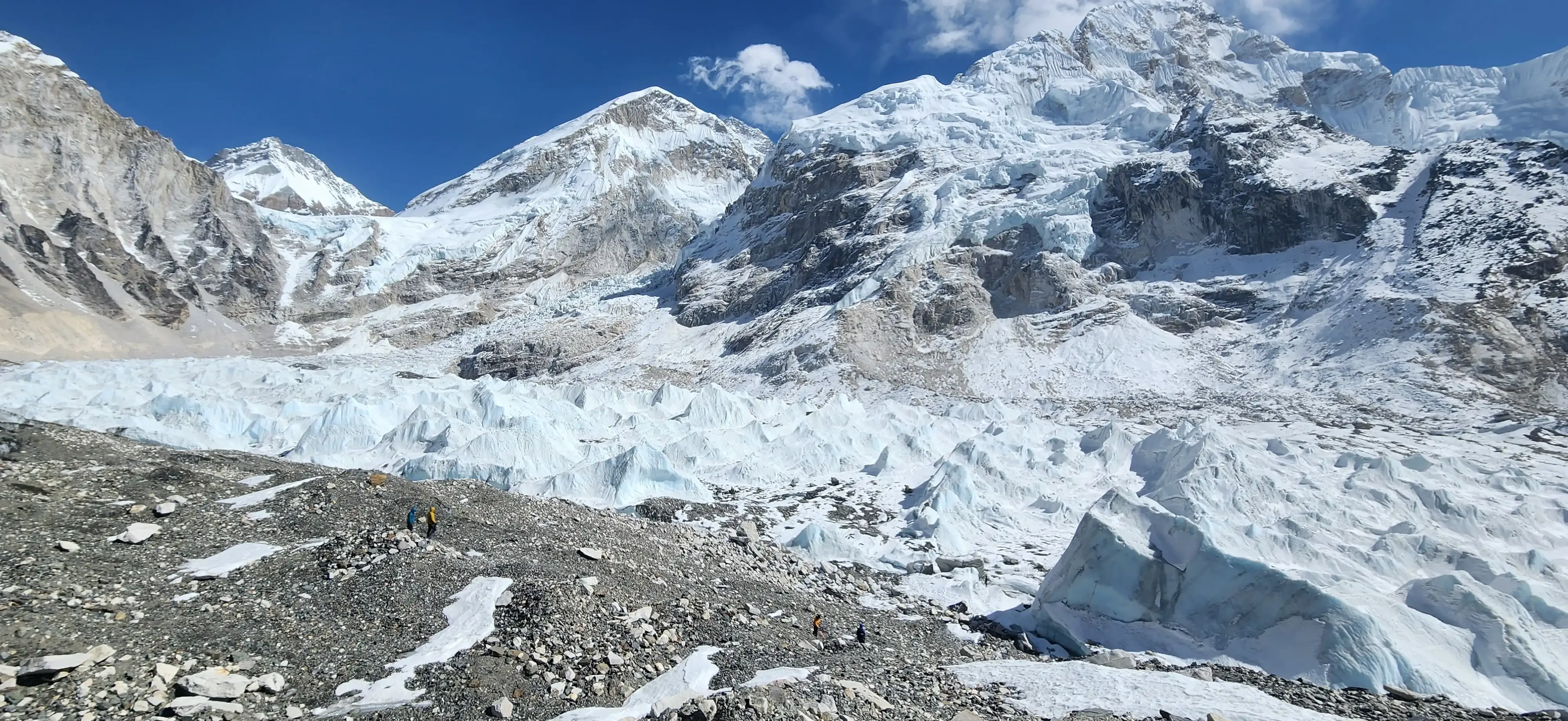 Trekkers cross rocky glacial terrain beneath towering snow-covered mountains and jagged ice formations.