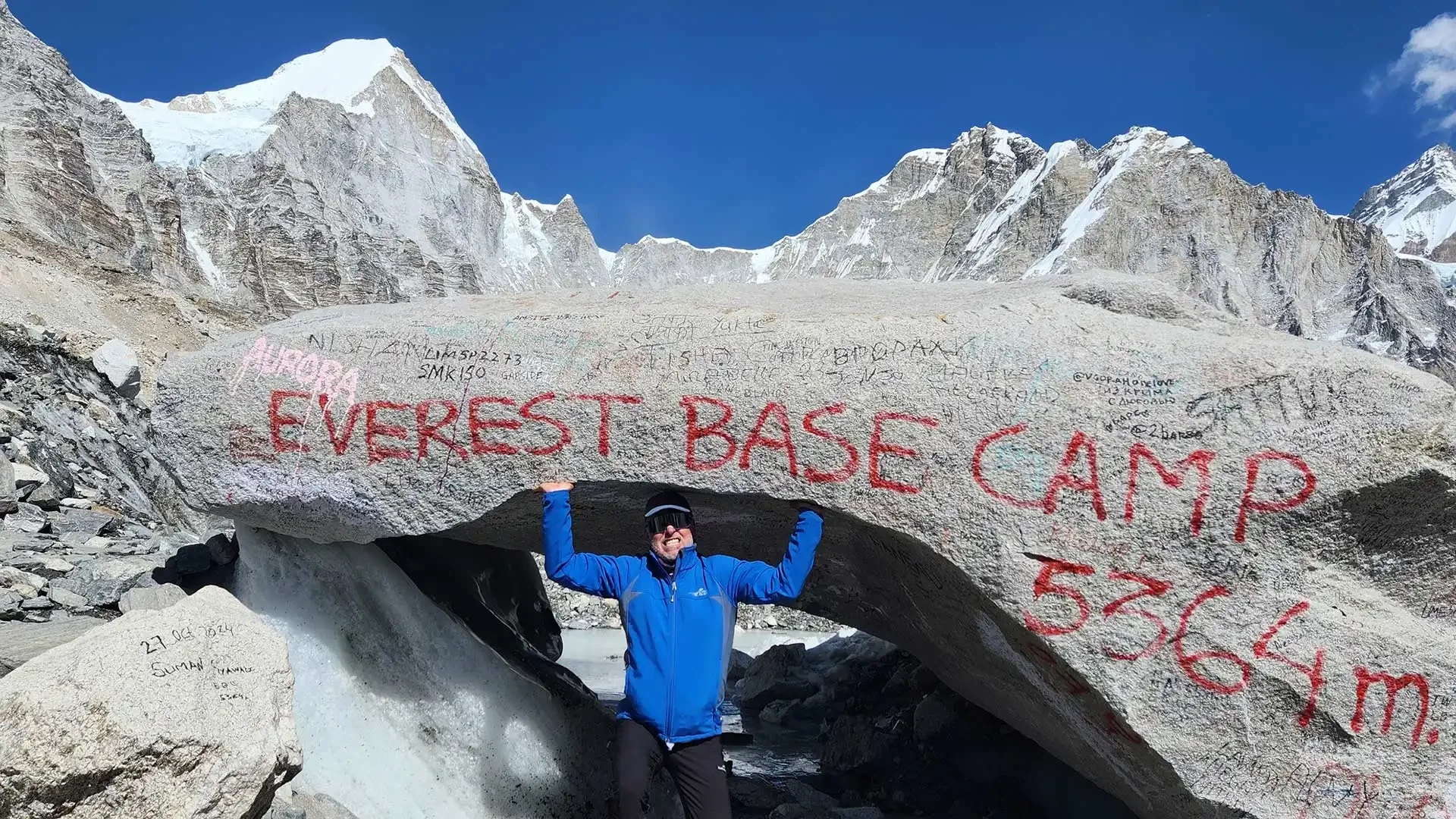 Trekker in blue jacket beneath Everest Base Camp marker boulder, surrounded by rugged snowy peaks.