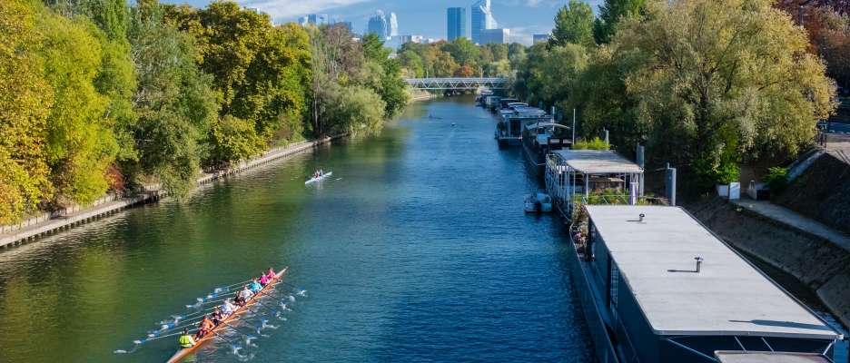 People rowing on the Seine, near Île de la Jatte, with barges and the La Défense district in the background