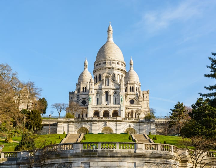 La basilique du Sacré-Cœur de Paris sous un ciel bleu printanier, vue depuis le bas du parc Louise Michel, avec la terrasse et les escaliers au premier plan.