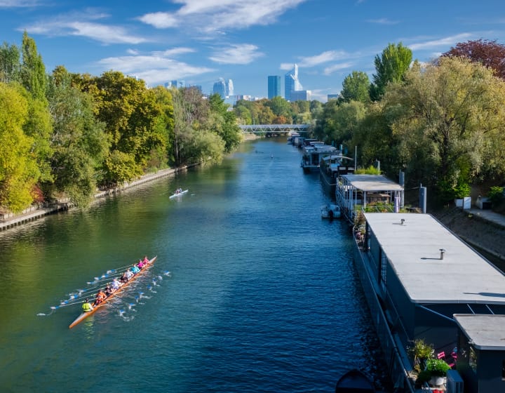 People rowing on the Seine, near Île de la Jatte, with barges and the La Défense district in the background