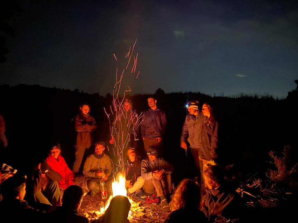 Mechina participants sitting on benches in a circle outdoors during sunset.