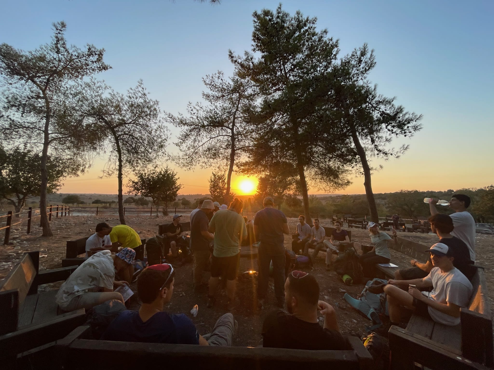 Mechina participants sitting on benches in a circle outdoors during sunset.