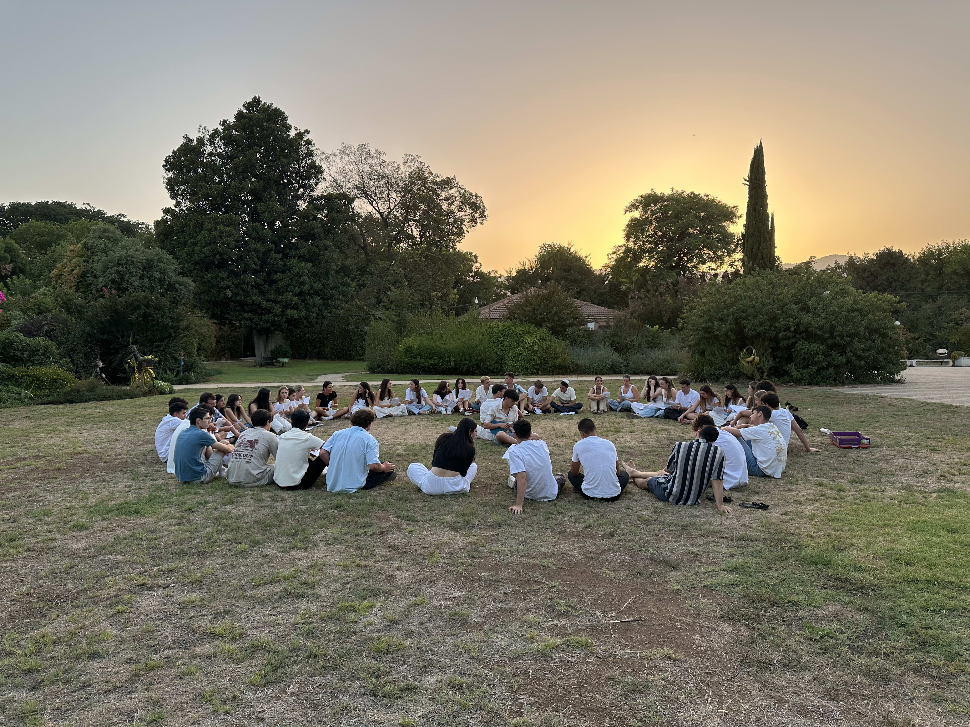 Participants sitting in a circle on grass in the sunset dressed in white for shabbat.