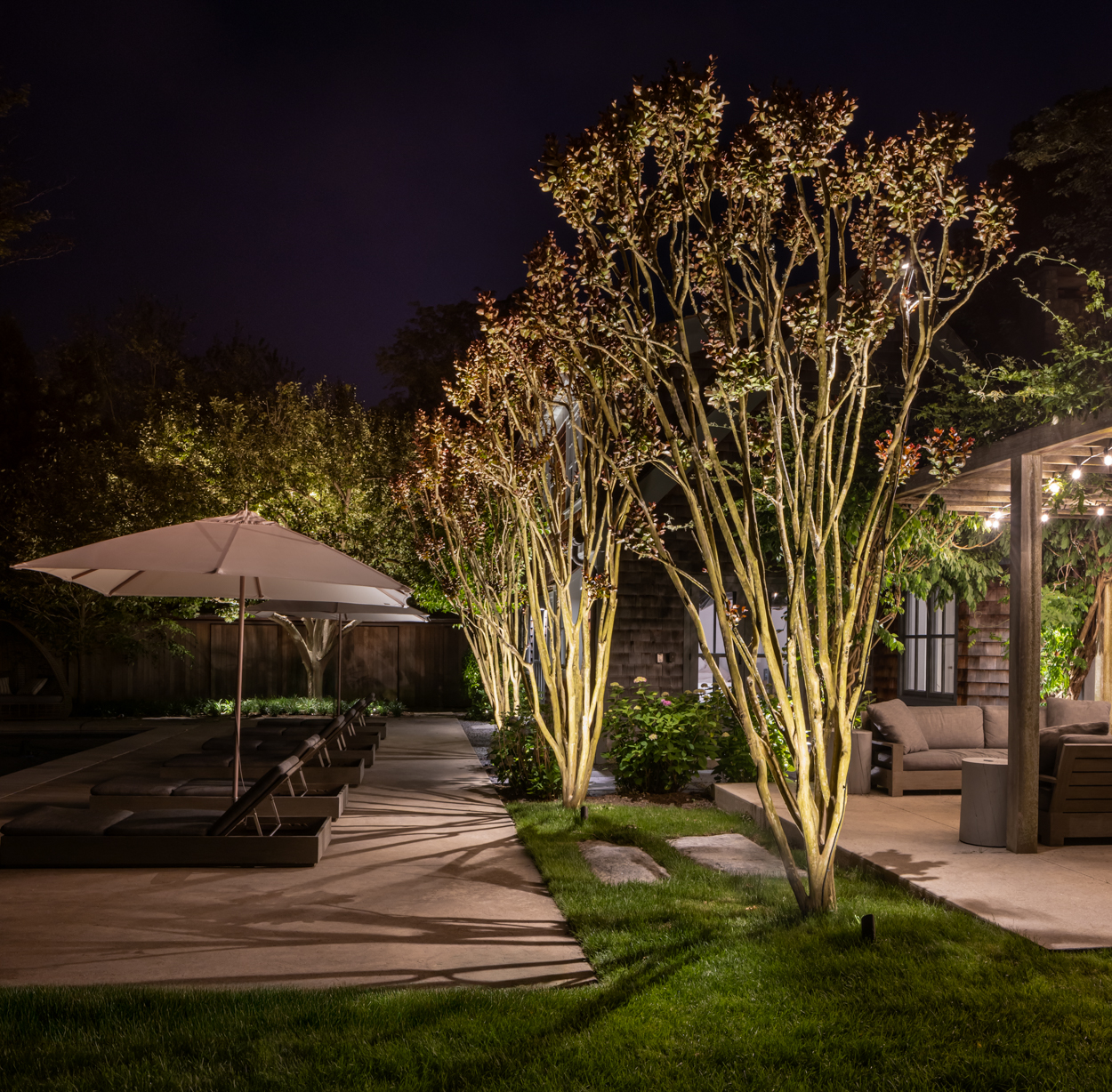 Landscape lighting adjusted around mature garden growth at a Hamptons estate — warm uplighting on hedges and planting beds at dusk