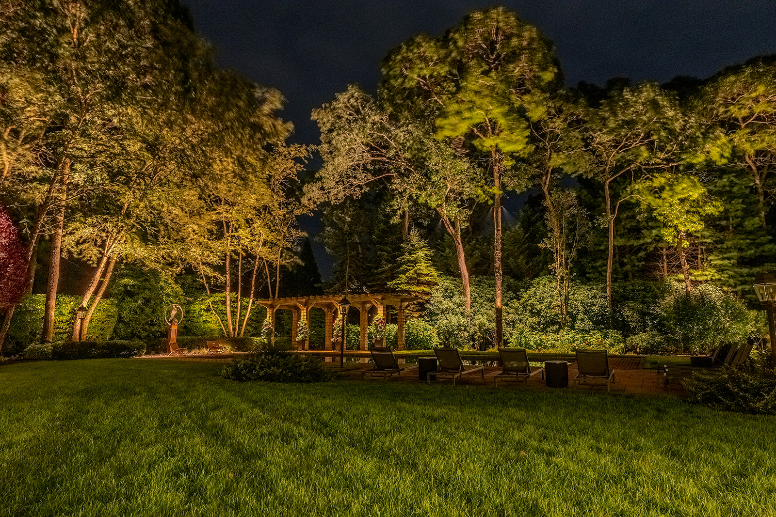 Wide nighttime view of illuminated trees, lawn, pool and pergola at a Quogue residence