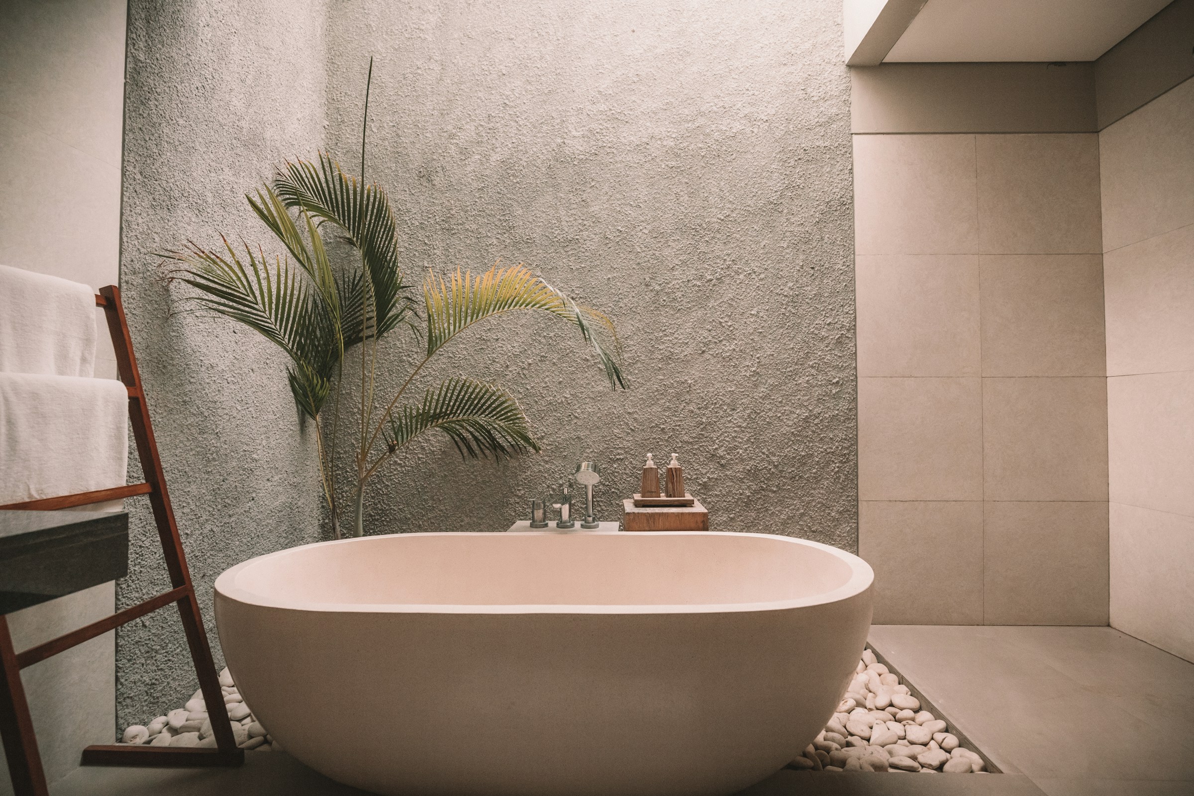 A modern bathroom showcases a sleek oval bathtub surrounded by white stones, exemplifying exquisite interior design services. A textured gray wall hosts a tall plant, while towels hang on a wooden rack and bath products neatly sit on a shelf beside the tub.