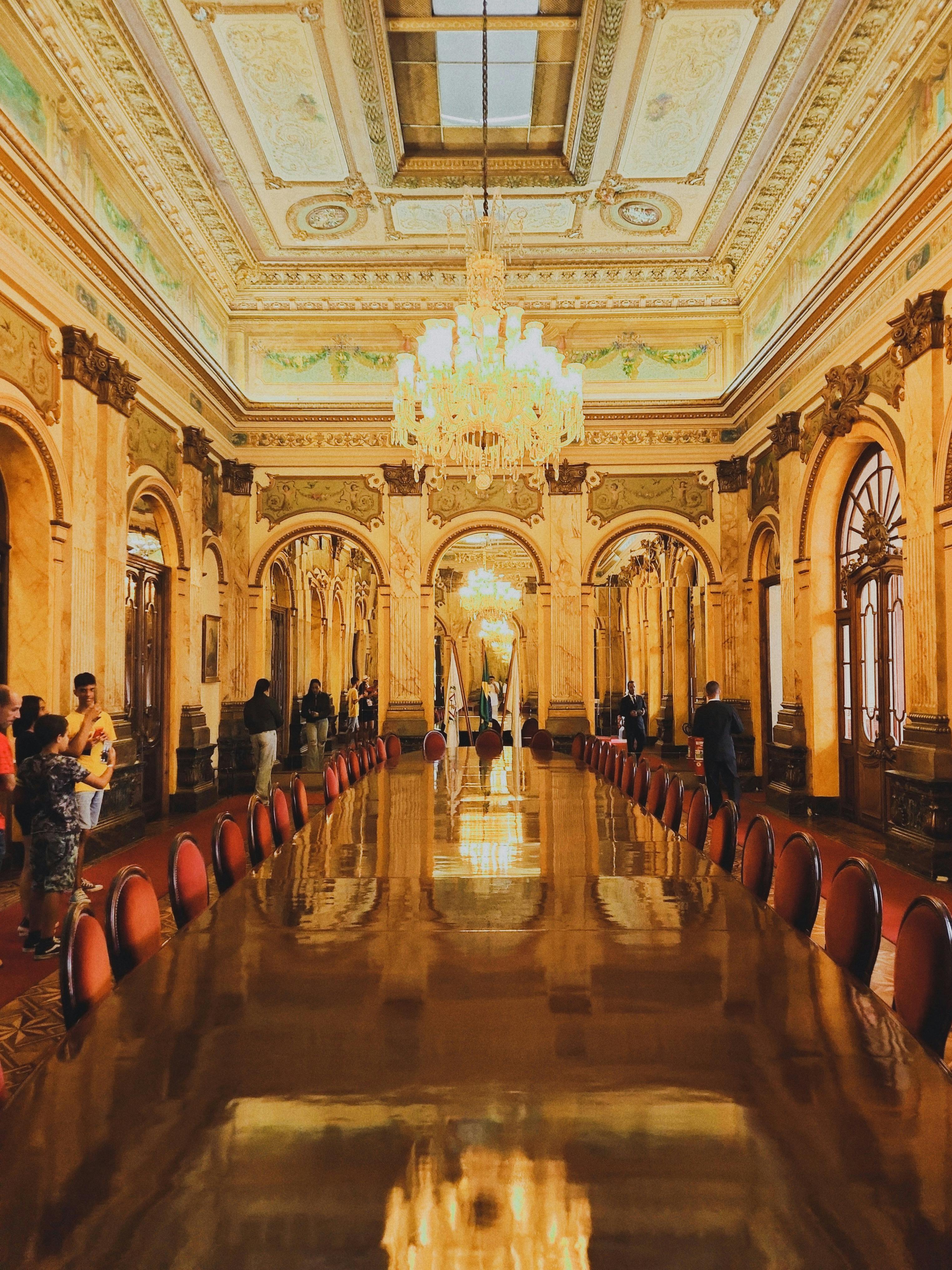 Elegant Interiors | Deco Serein A grand, opulent hall with ornate decor and high ceilings, an icon of exquisite interior design. Detailed frescoes and chandeliers adorn the space. A long, polished wooden table stretches through the center, surrounded by red upholstered chairs where people admire the elegant setting.
