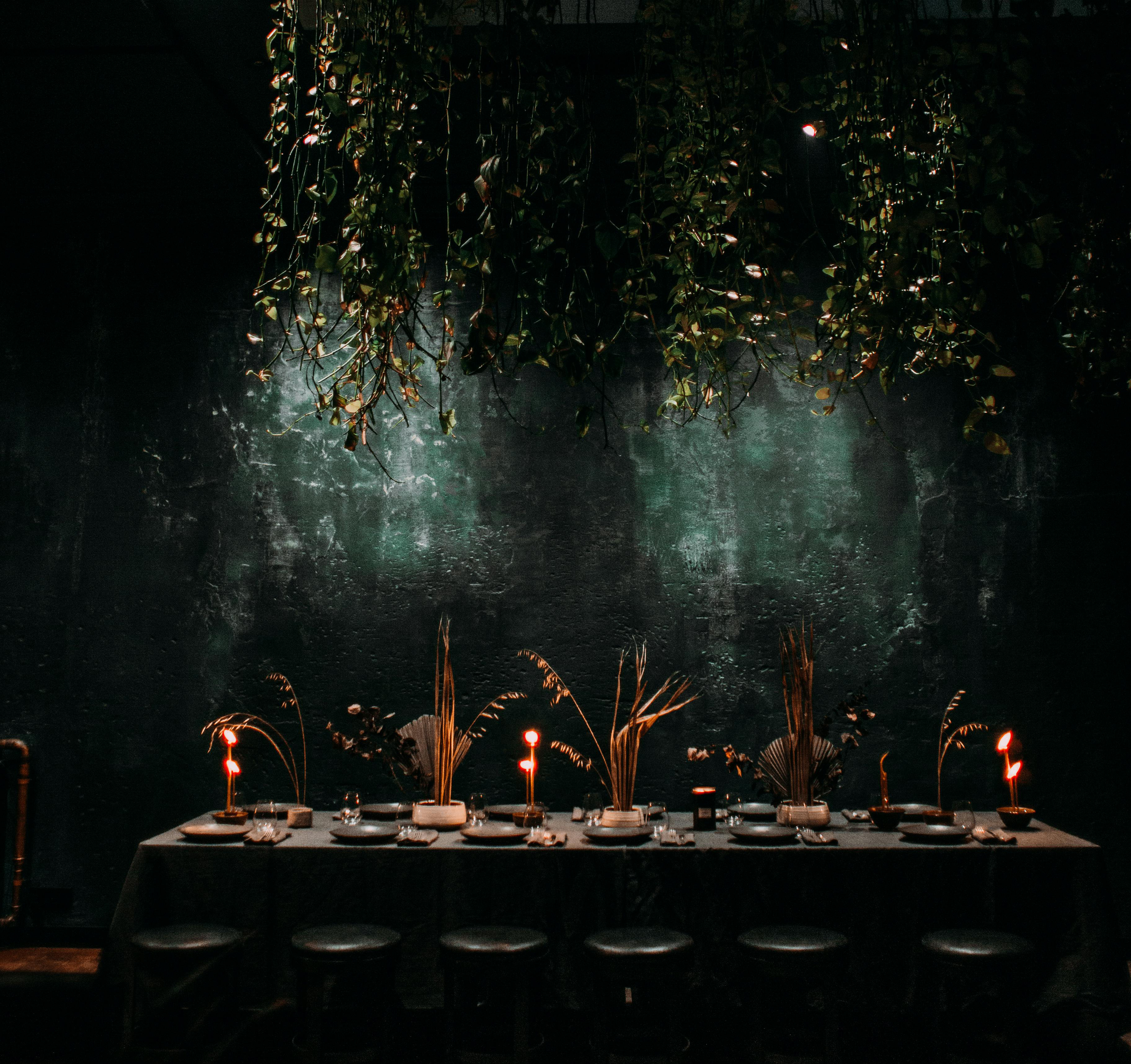 Theme-Based interior design | Deco Serein A dimly lit dining table with six chairs is elegantly set with candles, plates, and decorative plants. Green foliage hangs from above, set against a dark, textured wall, creating an intimate and atmospheric home space that showcases impeccable interior design.