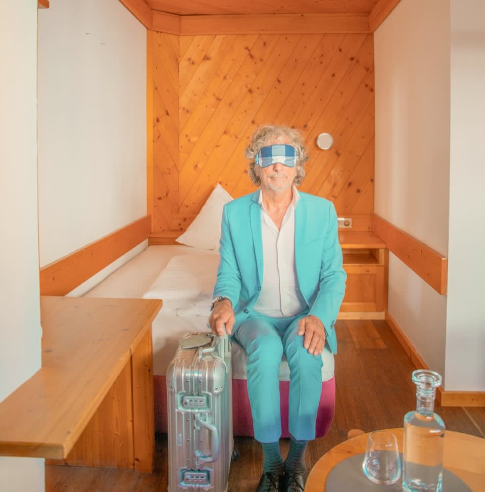 A man in a blue suit sits with a suitcase in a wooden room, possibly at Glemmtalerhof in Hinterglemm.